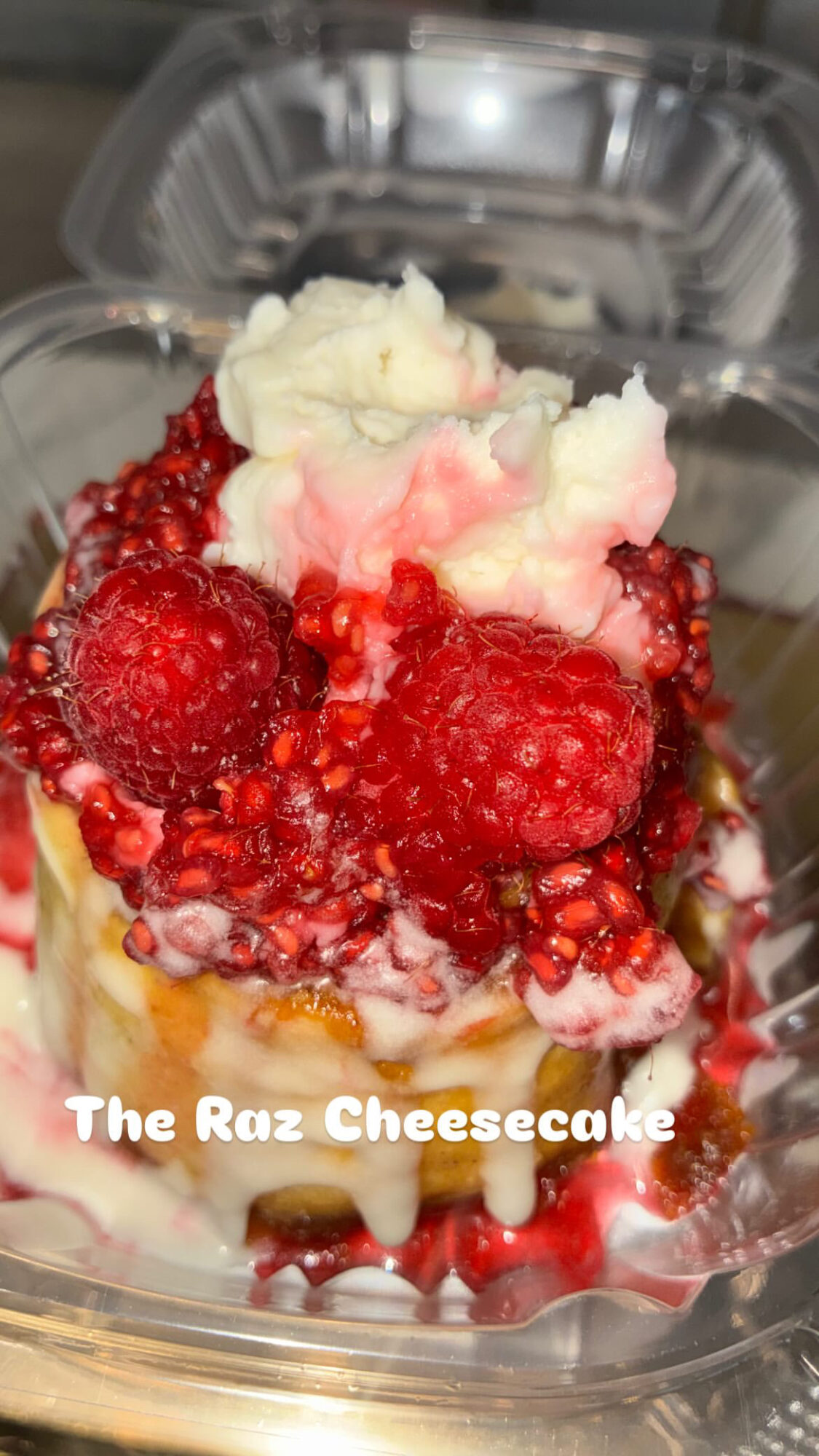 Cheesecake topped with whipped cream, raspberries, and red fruit sauce in a clear container.