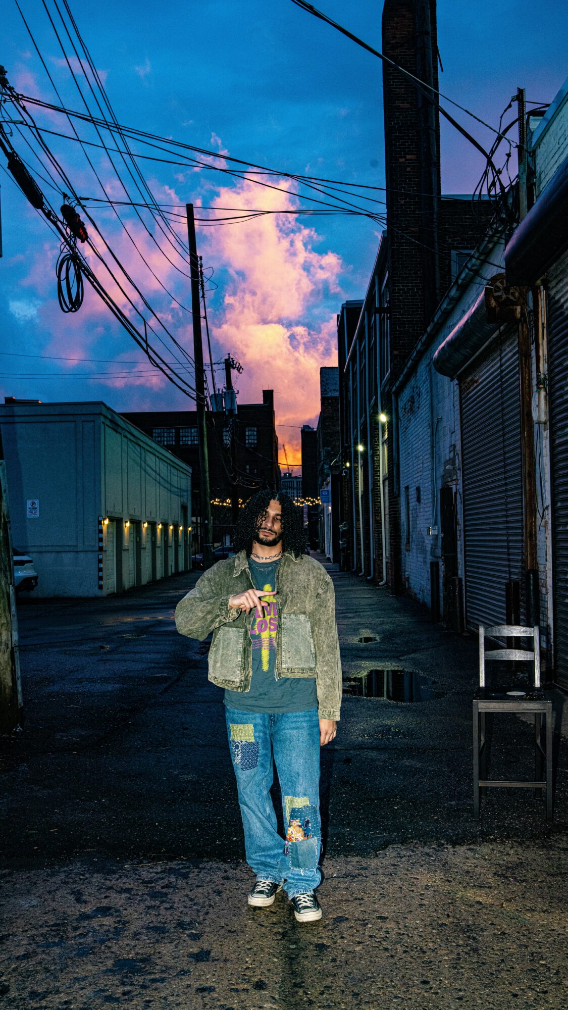 Person standing in an alleyway with colorful sky and clouds, wearing a jacket and jeans, holding a drink.