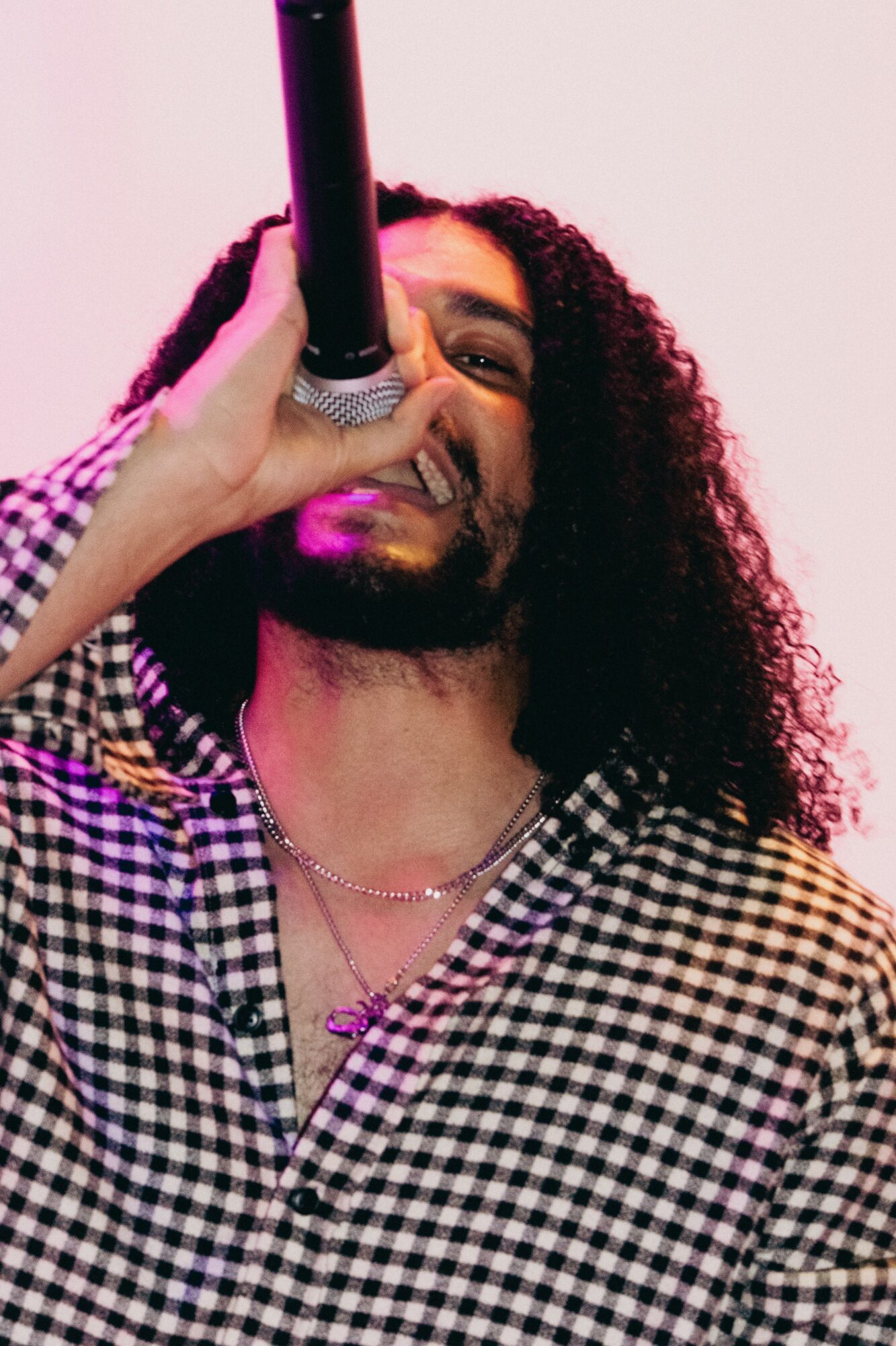 Person with long curly hair singing into a microphone, wearing a checkered shirt and necklace, against a pink background.