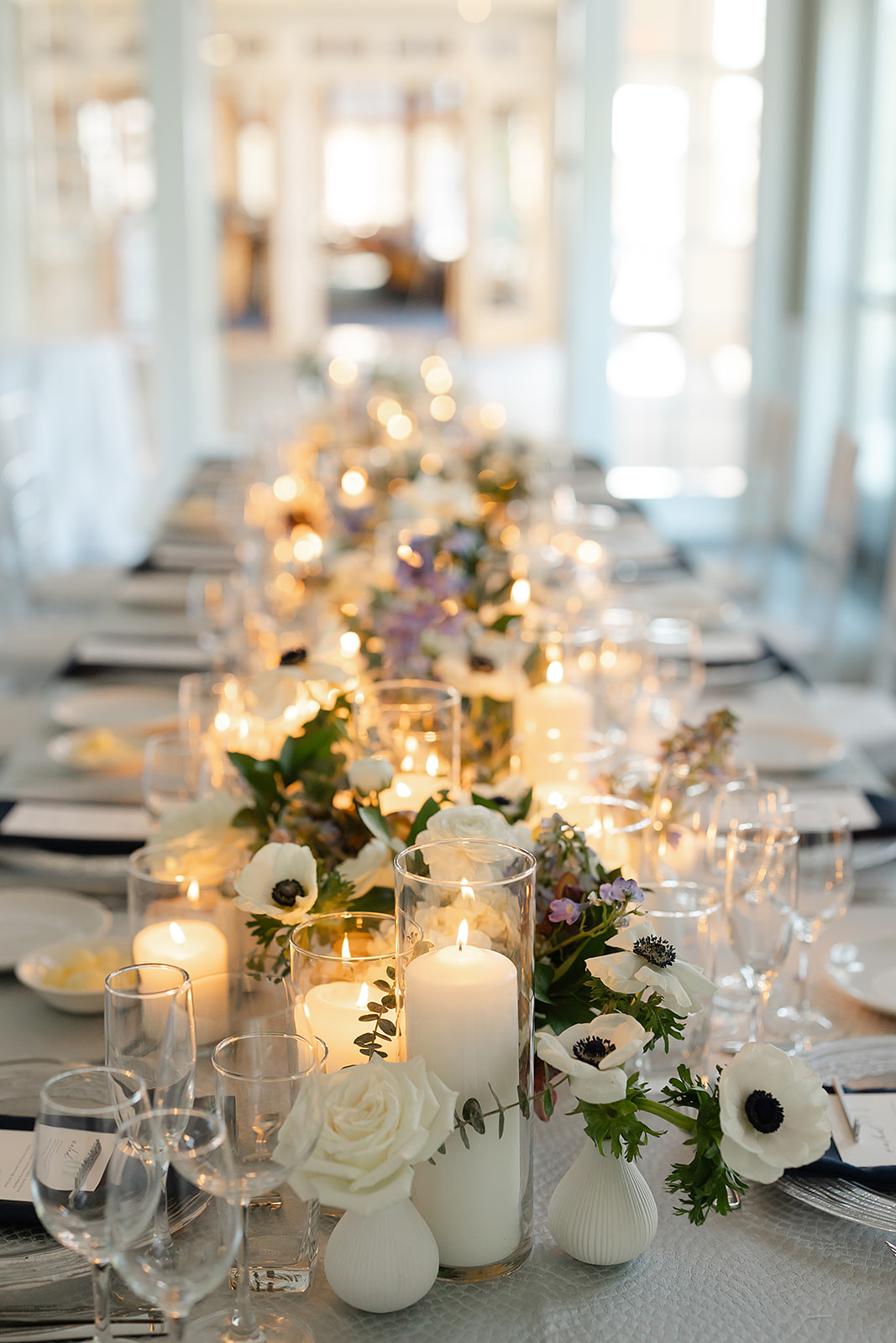 Long dining table decorated with candles, flowers, and greenery in a bright, airy room with large windows.