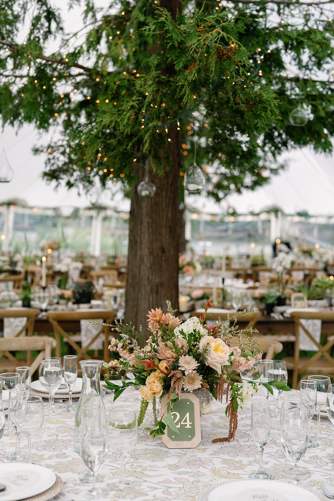 Table with floral centerpiece and a green table number card, set outdoors with a large tree in the background.