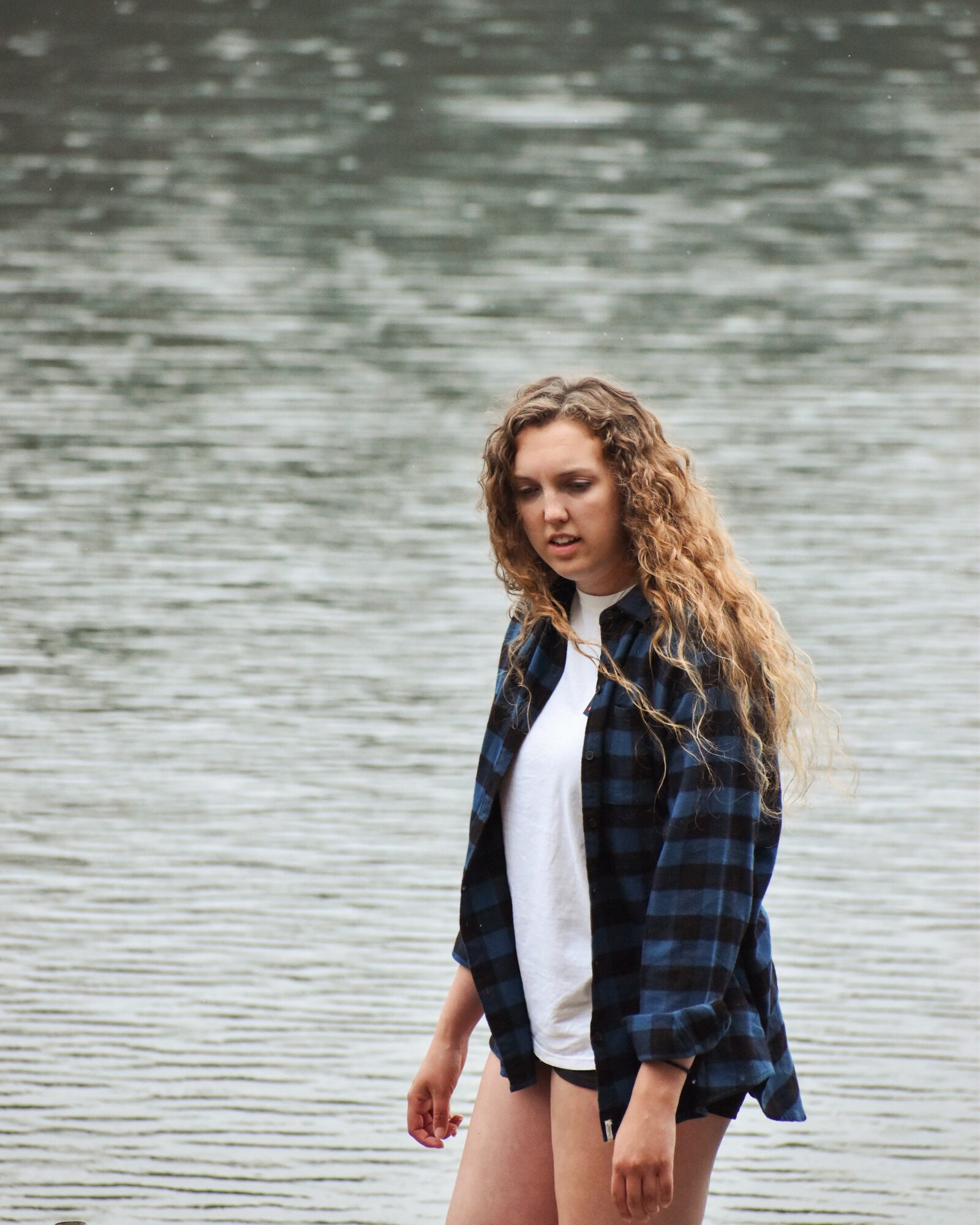 Young woman with curly hair standing near water, wearing a plaid shirt over a white top, with a neutral expression.