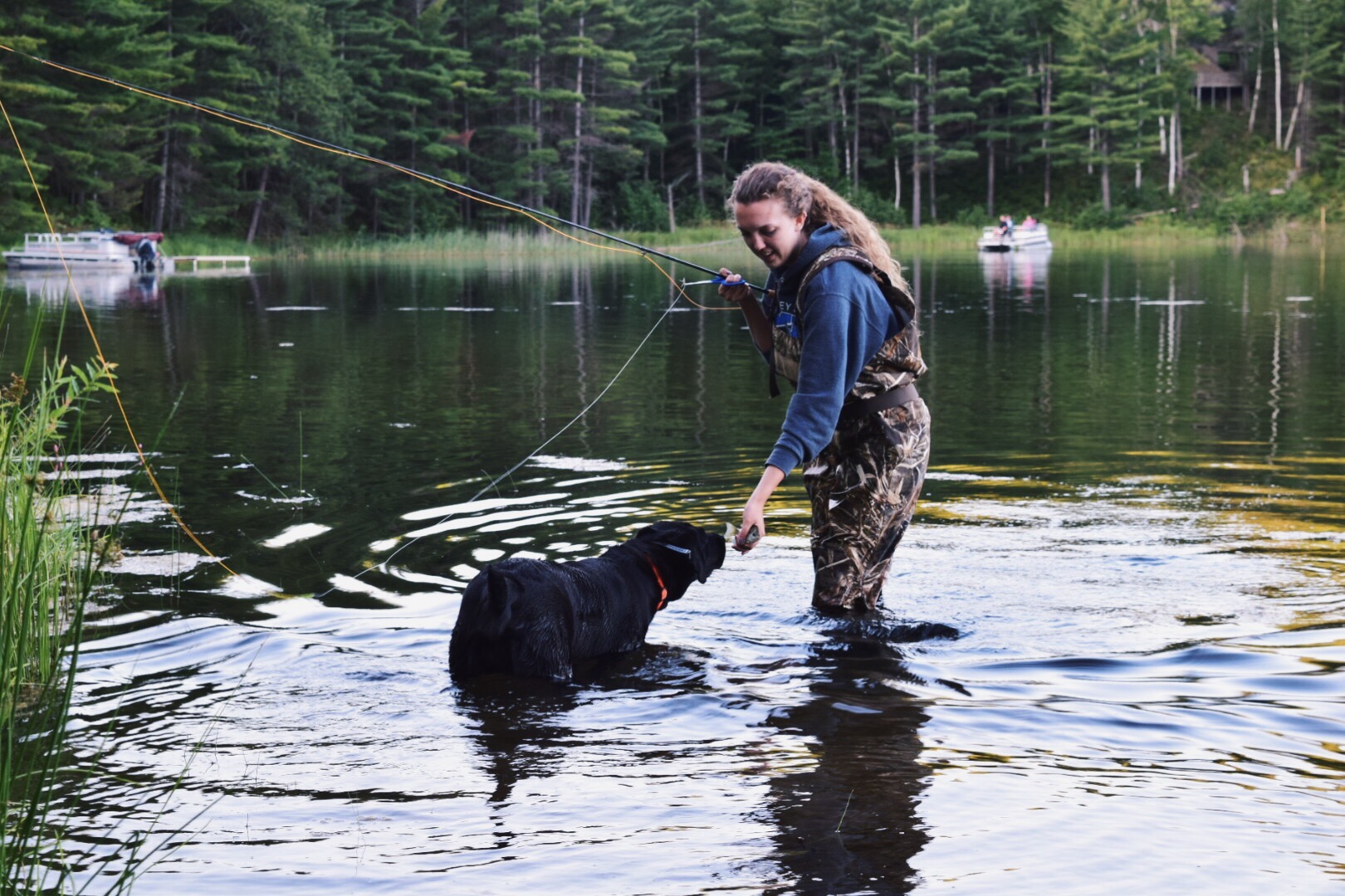 Person standing in water with a dog, fishing in a lake surrounded by trees, calm water, and a forest background.