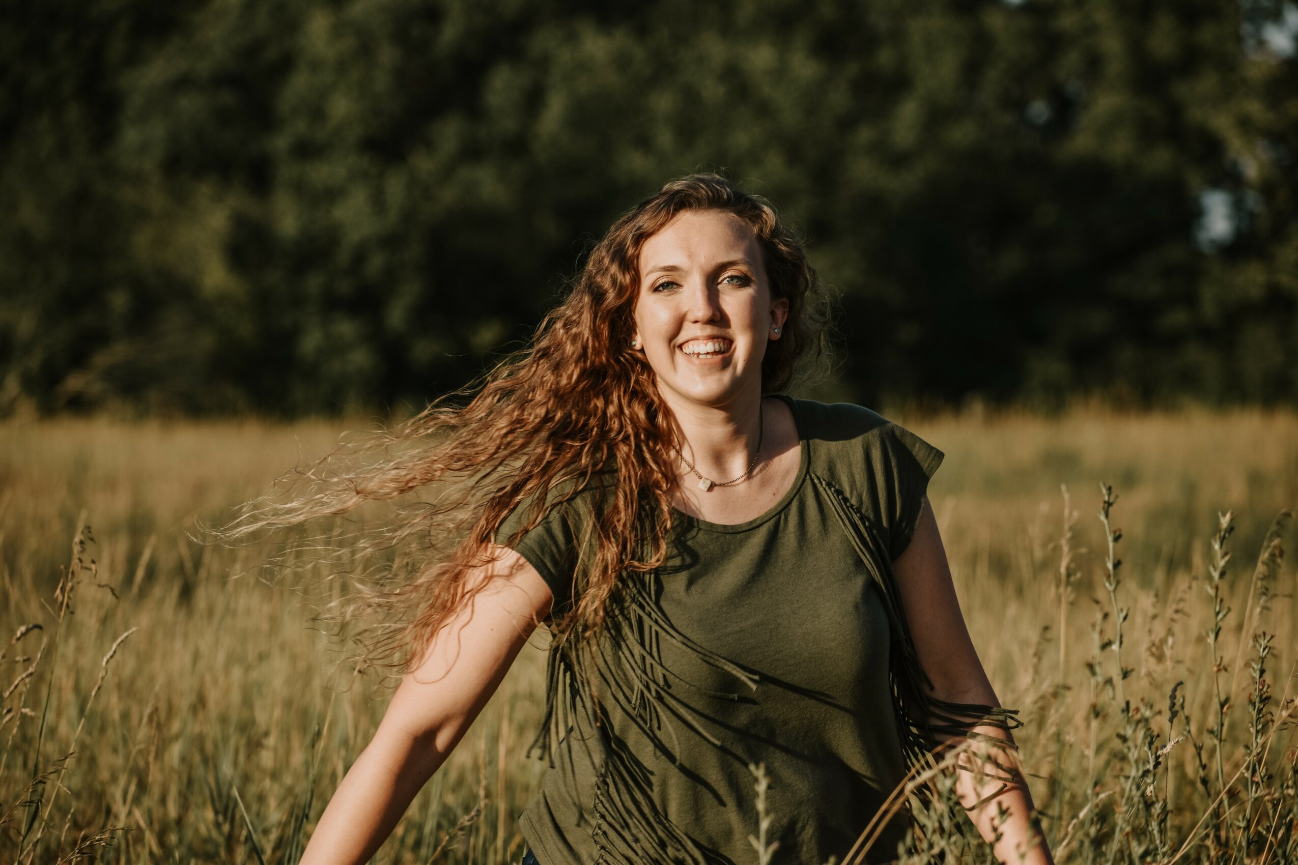 Smiling woman with long curly hair standing in a grassy field, trees in the background.