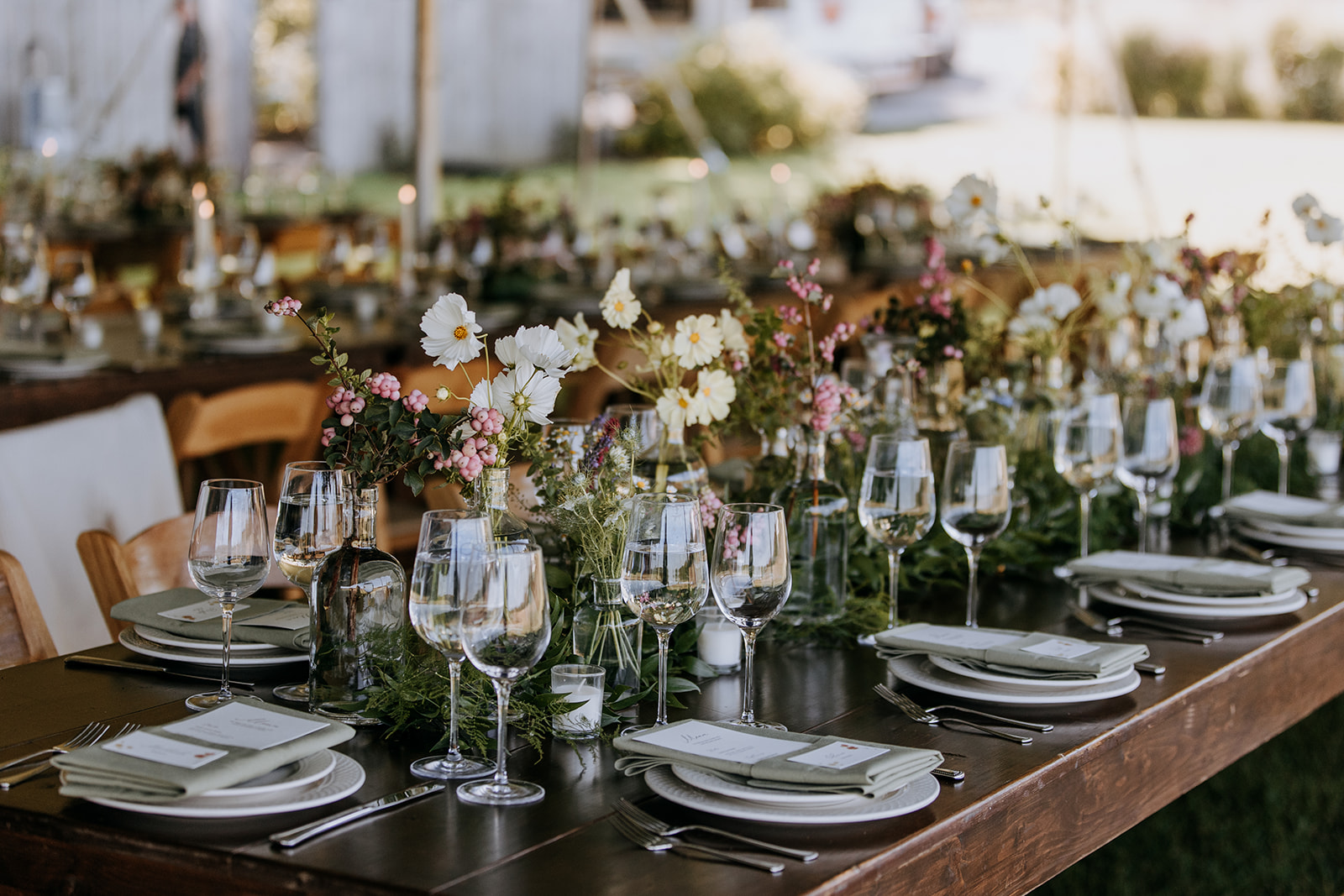 Long table set with plates, glasses, and floral centerpieces outdoors, with chairs and a blurred background.