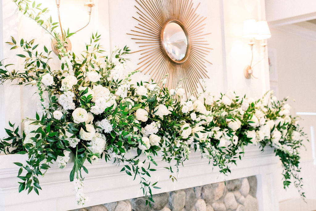 Large floral arrangement with white flowers and green foliage on a white fireplace mantel, with a sunburst mirror and wall sconce above.
