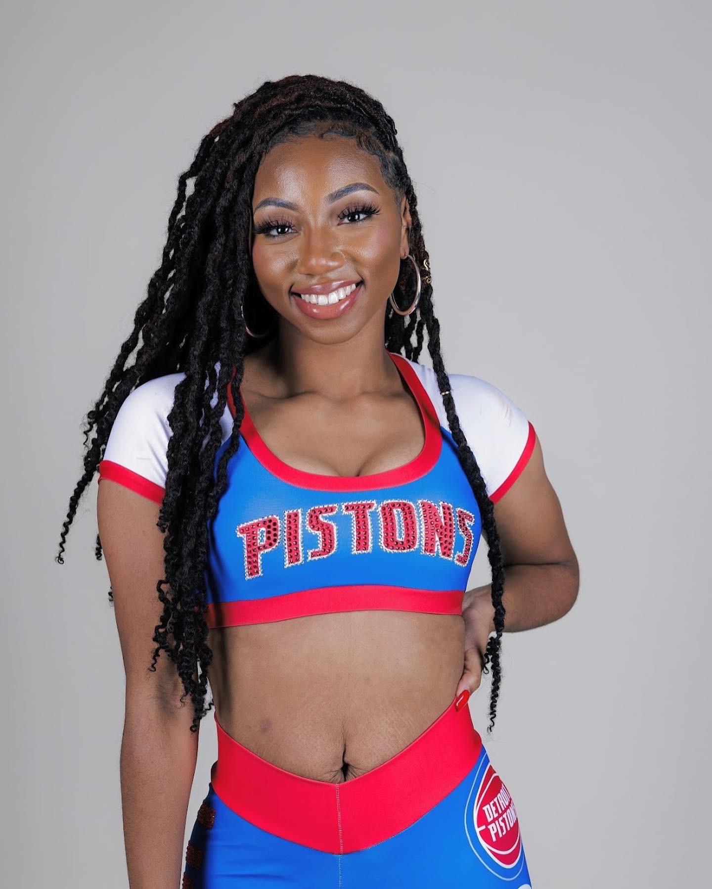 Smiling woman with long dreadlocks wearing a Detroit Pistons cheerleading outfit, standing against a plain background.