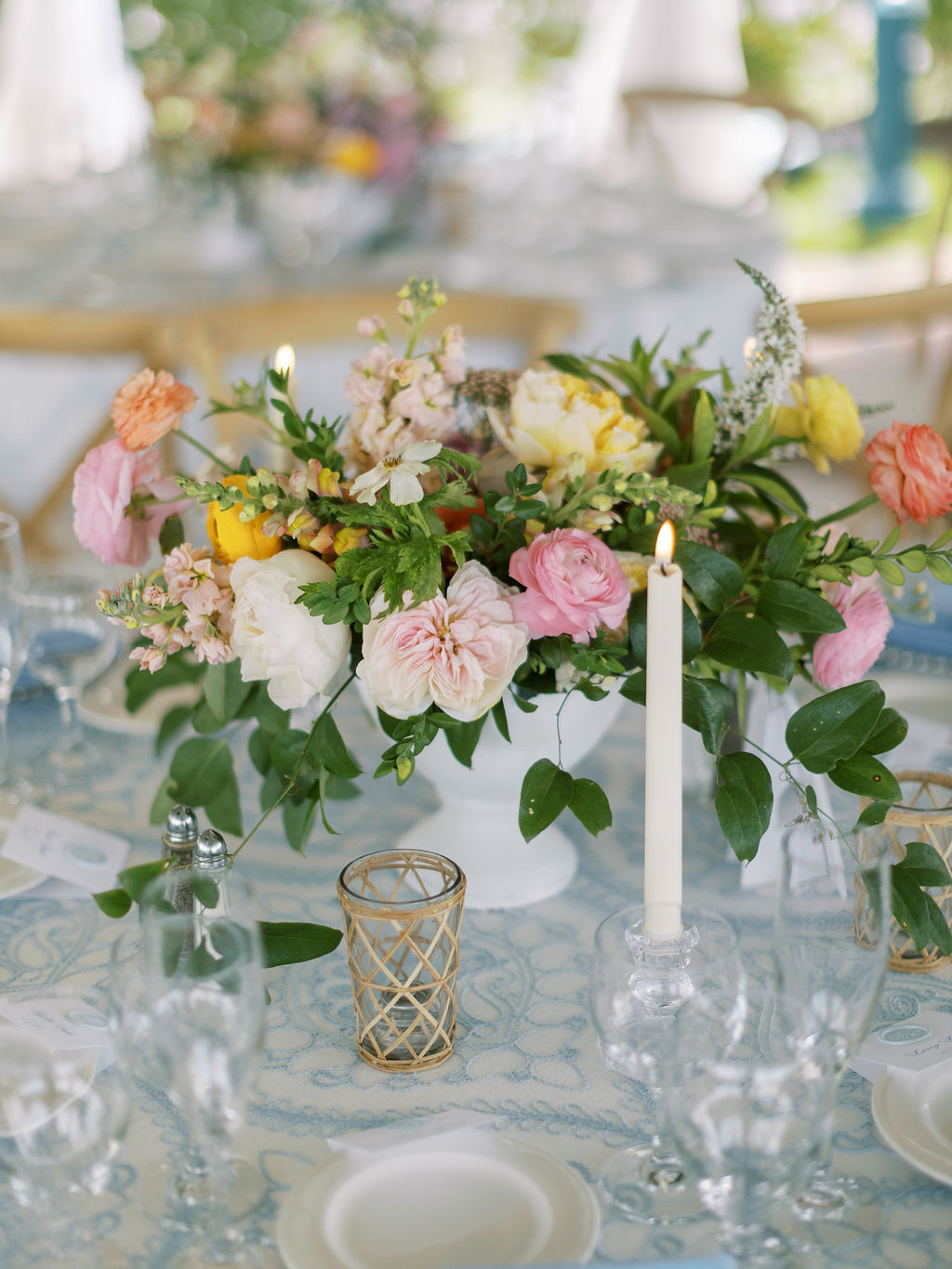 Floral centerpiece with pink, white, and yellow flowers, green leaves, and candles on a table.