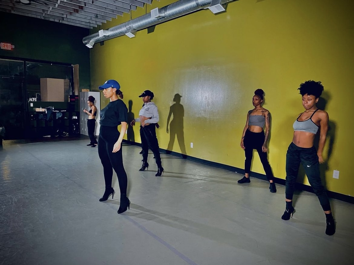 Five women in dance practice in a studio with a yellow wall, some wearing crop tops and jeans, others in casual attire.