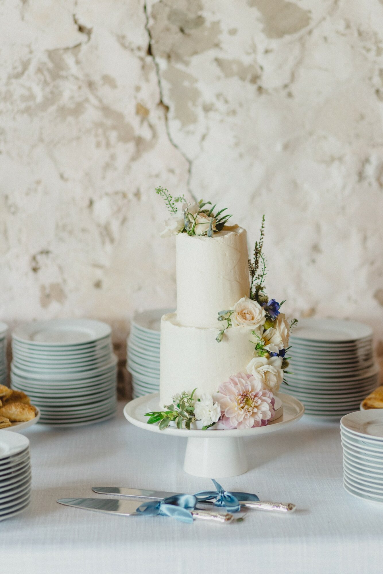 Two-tiered white cake decorated with flowers on a table, surrounded by stacks of plates and a knife with a blue ribbon.