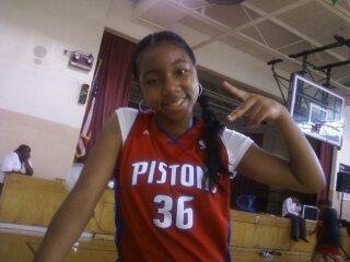 Girl in a red basketball jersey smiling and pointing to her head in a gymnasium.