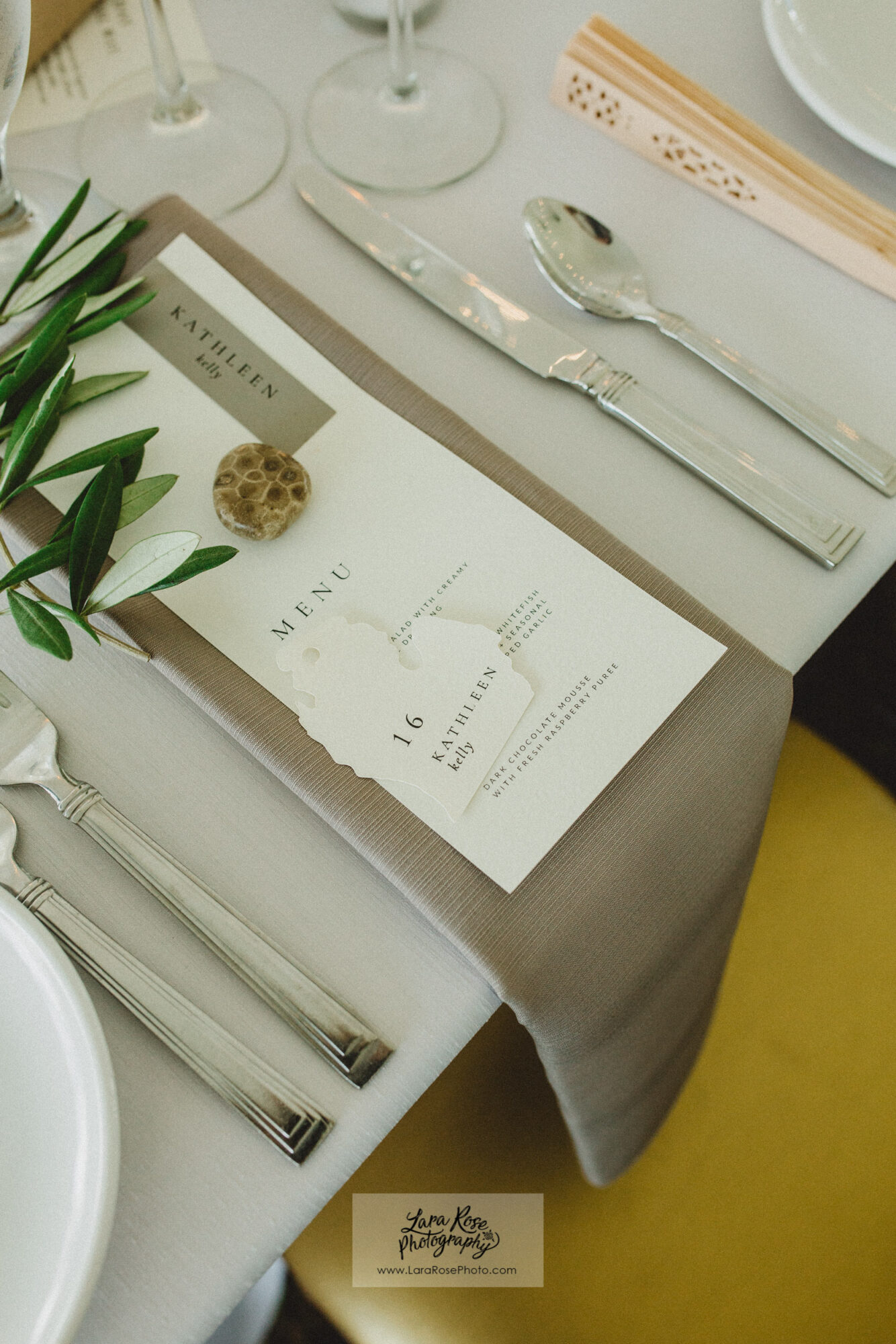 Table setting with menu, spoon, and napkin, decorated with greenery and a small flower, on a beige tablecloth.