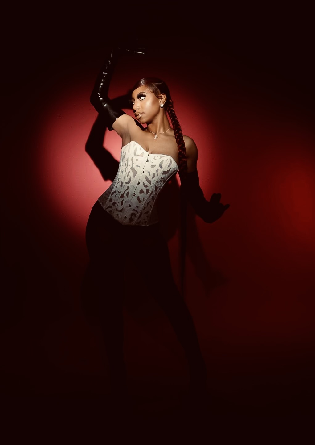 Woman in a corset and gloves poses against a red background, with one arm raised and head turned sideways.