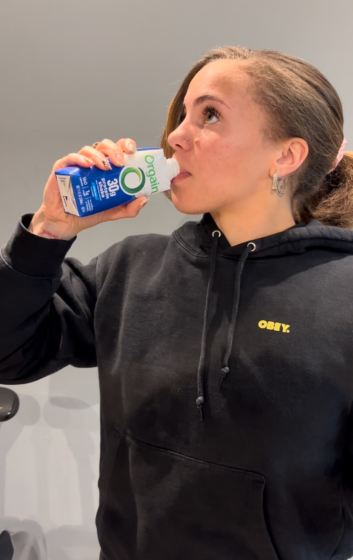 Young woman drinking from a blue carton, wearing a black hoodie with yellow text, against a plain background.