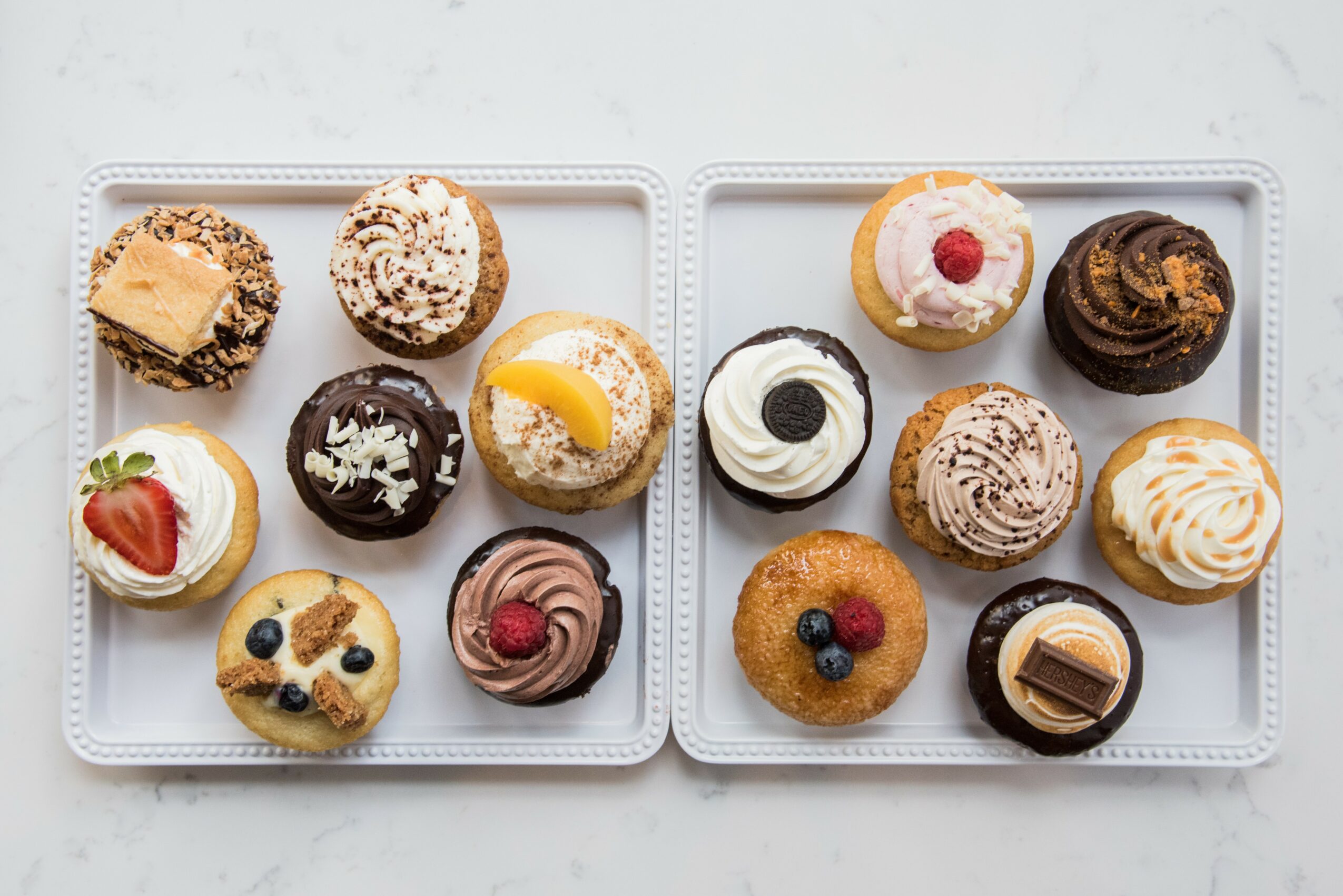 Two white trays with assorted cupcakes and muffins on a white surface, some topped with fruit or frosting.