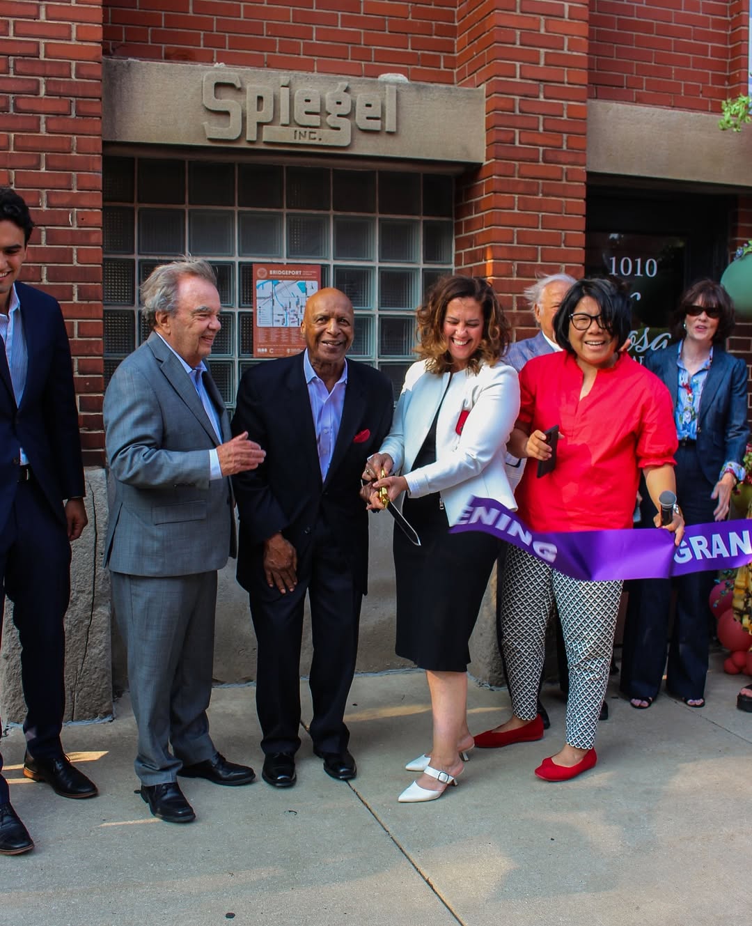Group of people at ribbon-cutting event outside brick building with sign reading 'Splage'.