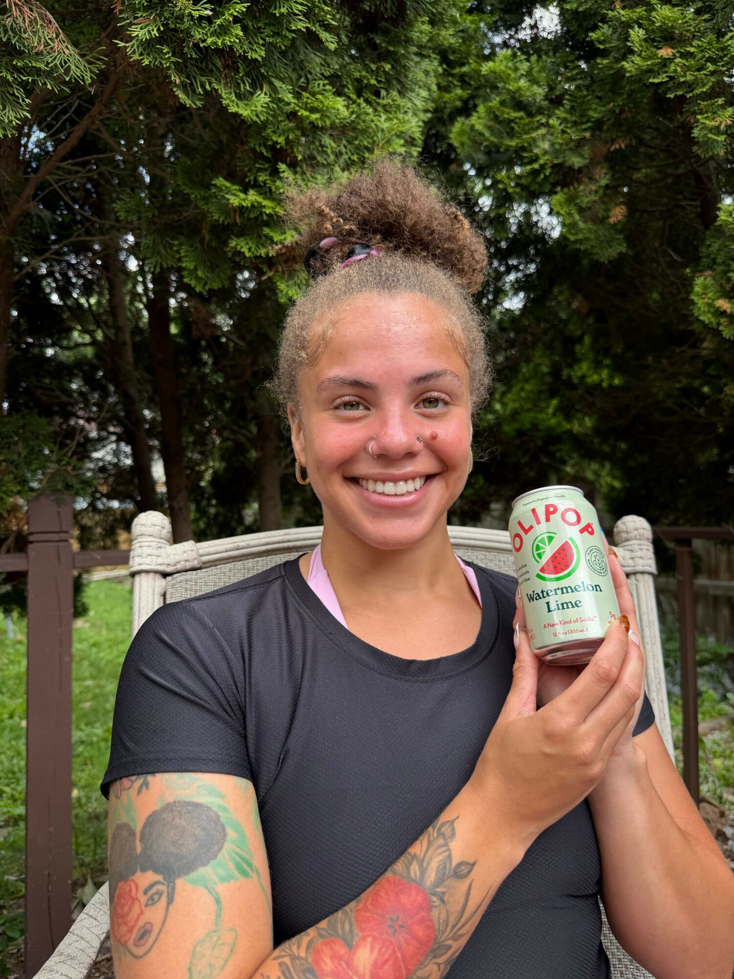 Woman with tattoos smiling holding a can outdoors with trees in background.