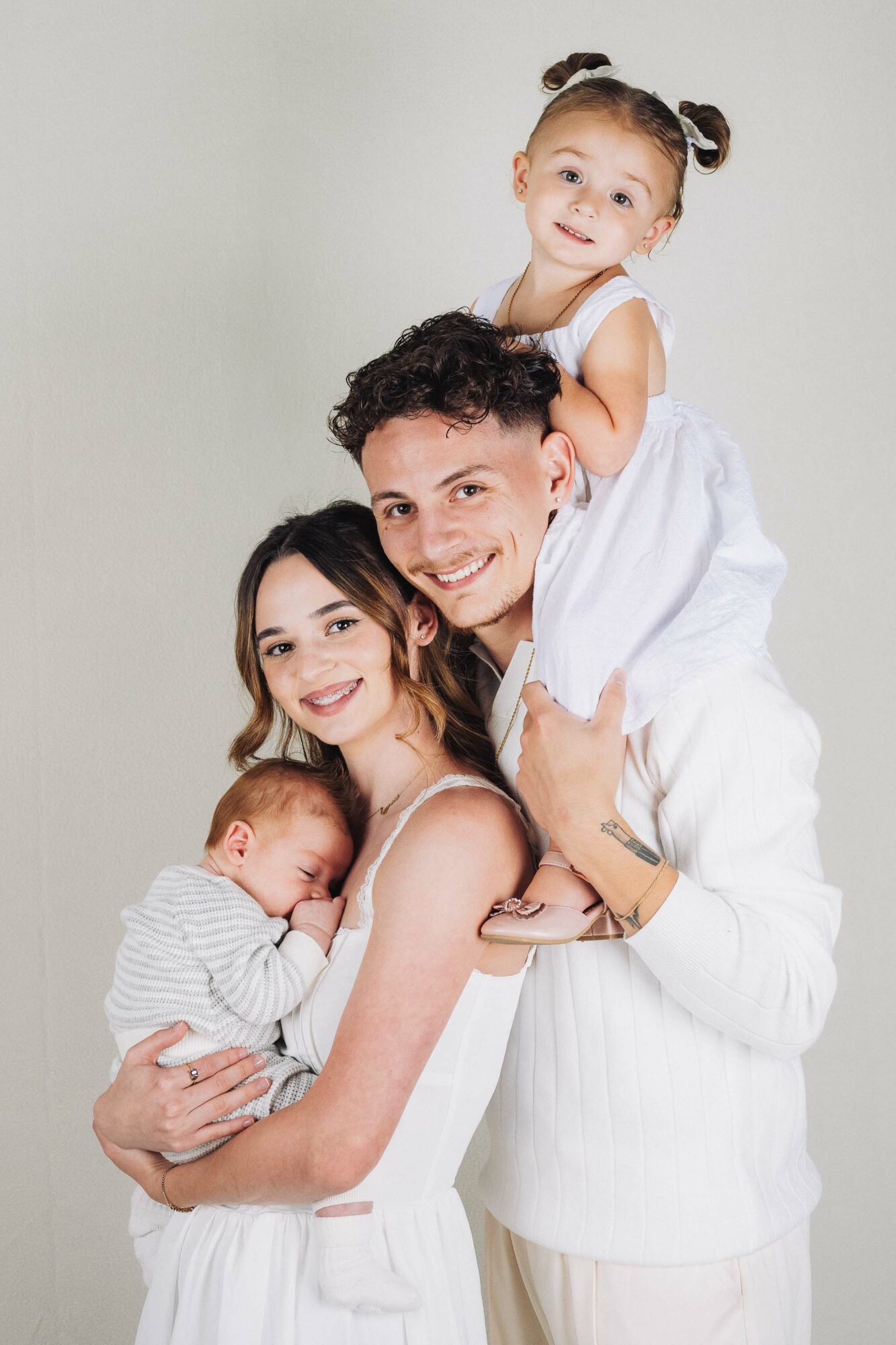 Family of four with a young girl on father's shoulders, mother holding a baby, all smiling against a plain background.