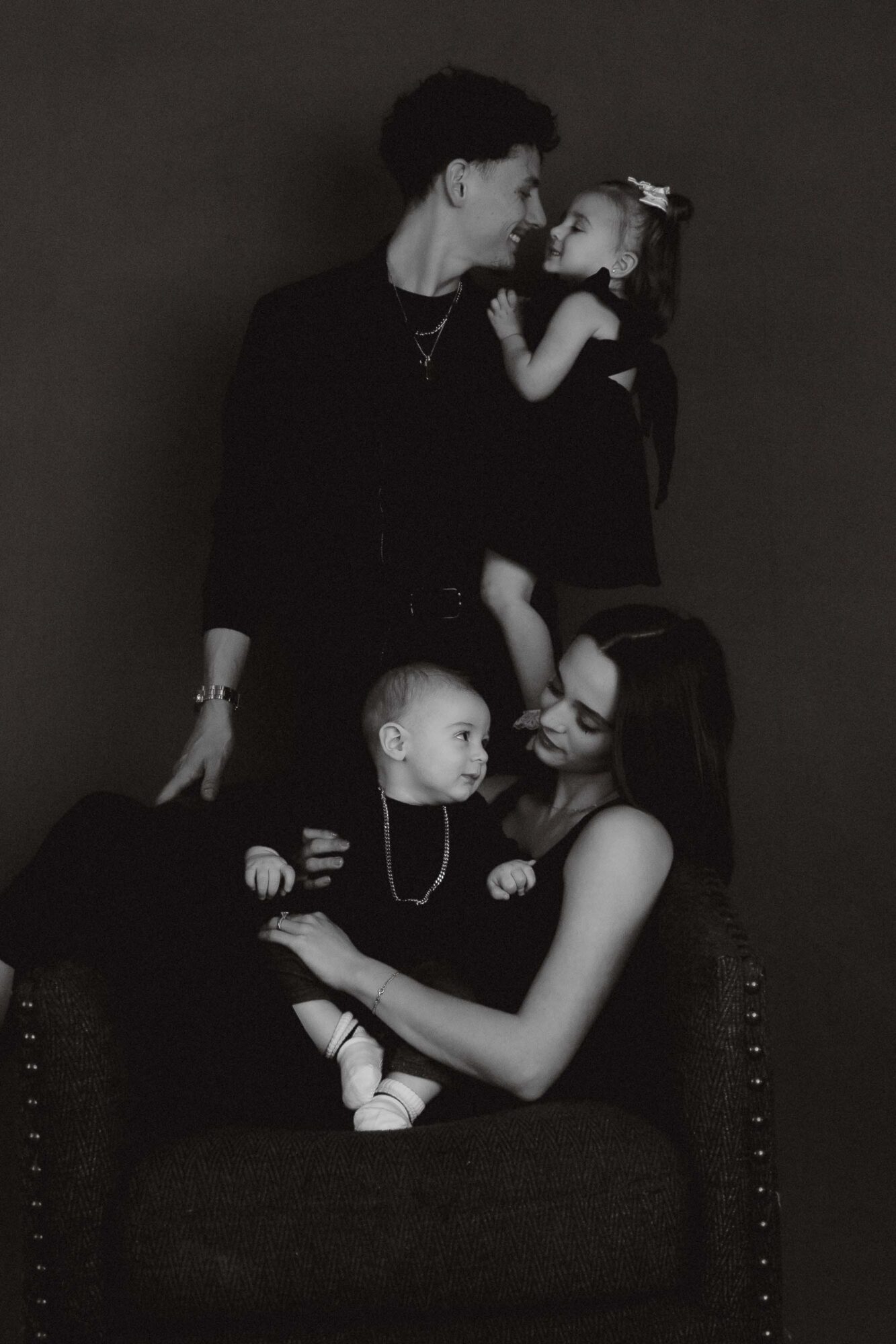 A woman with two children, one on her lap and one standing, all facing each other, in a dark setting.