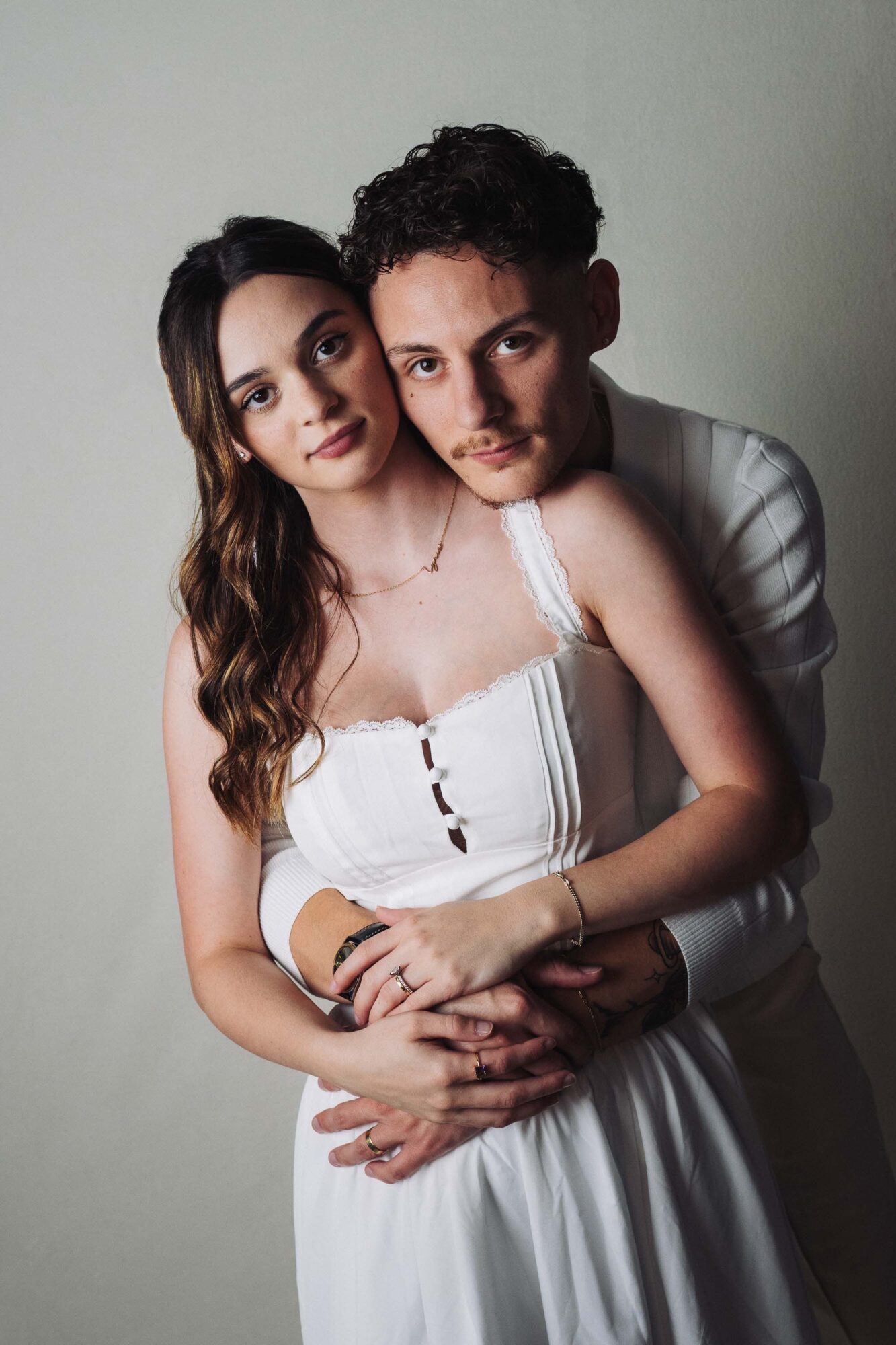 A young couple stands close together, the woman in a white dress and the man behind her, both looking at the camera.