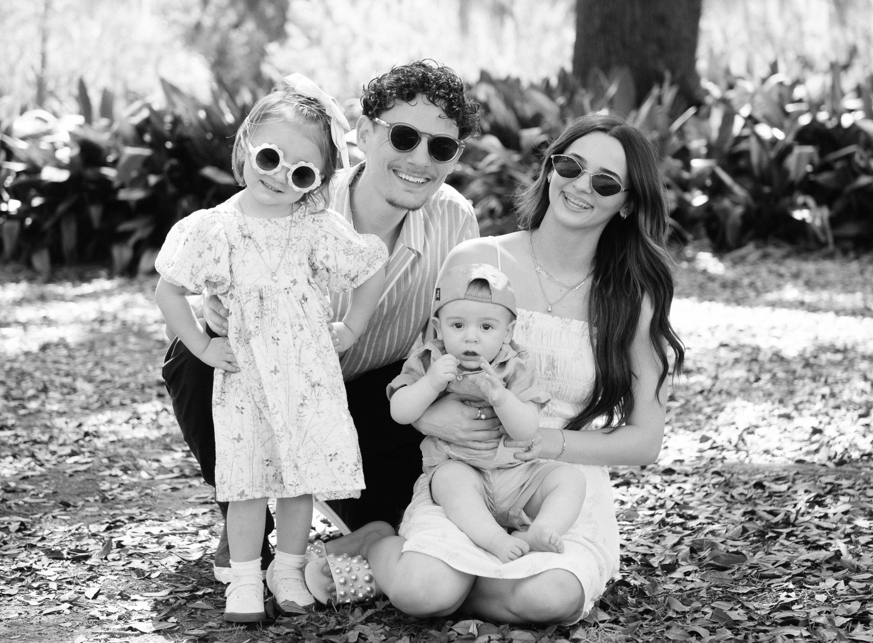 Family of four outdoors, two children and two adults, sitting on ground with trees and plants in background.