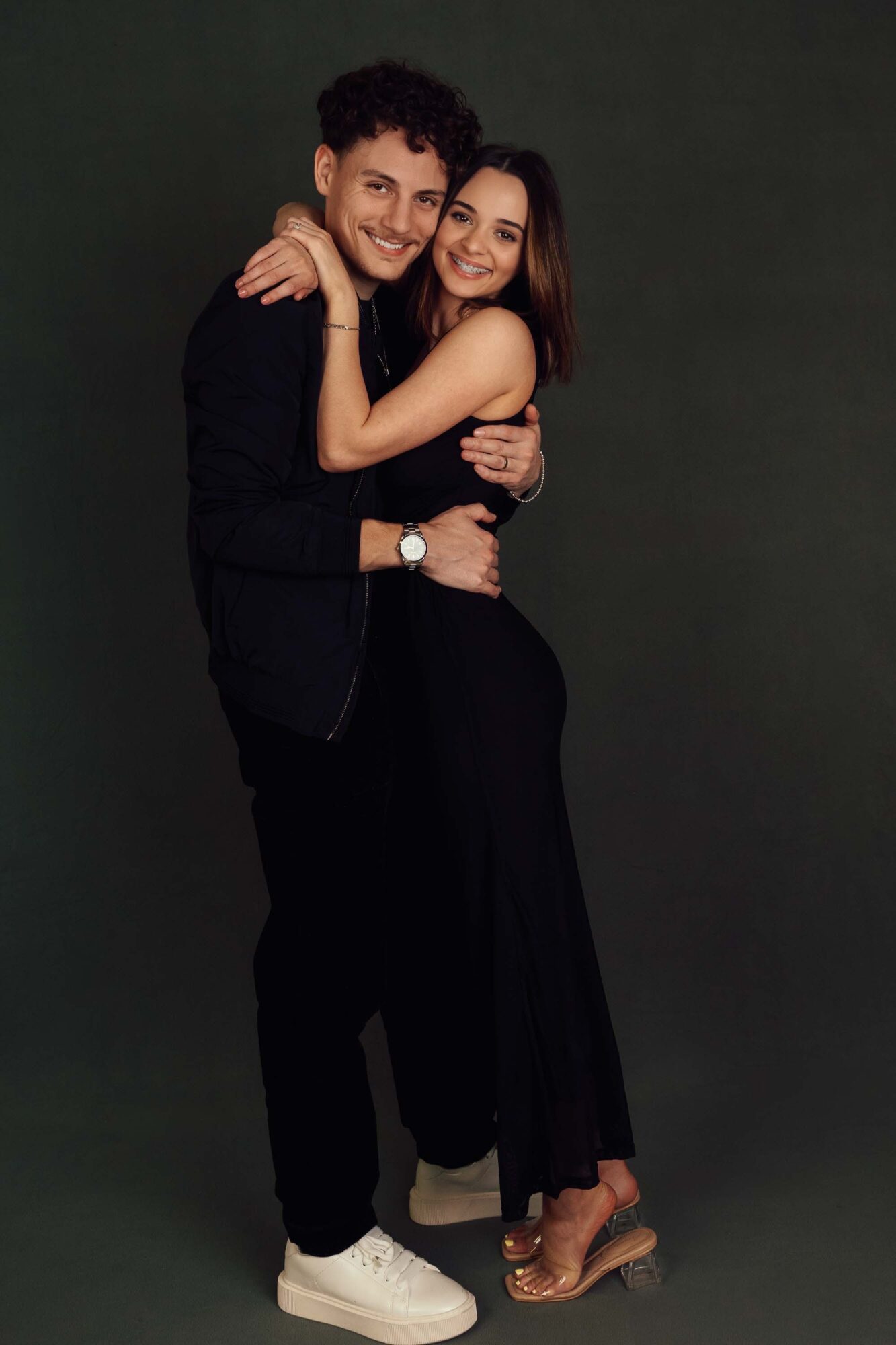 A young man and woman embrace and smile, standing close together against a dark background.