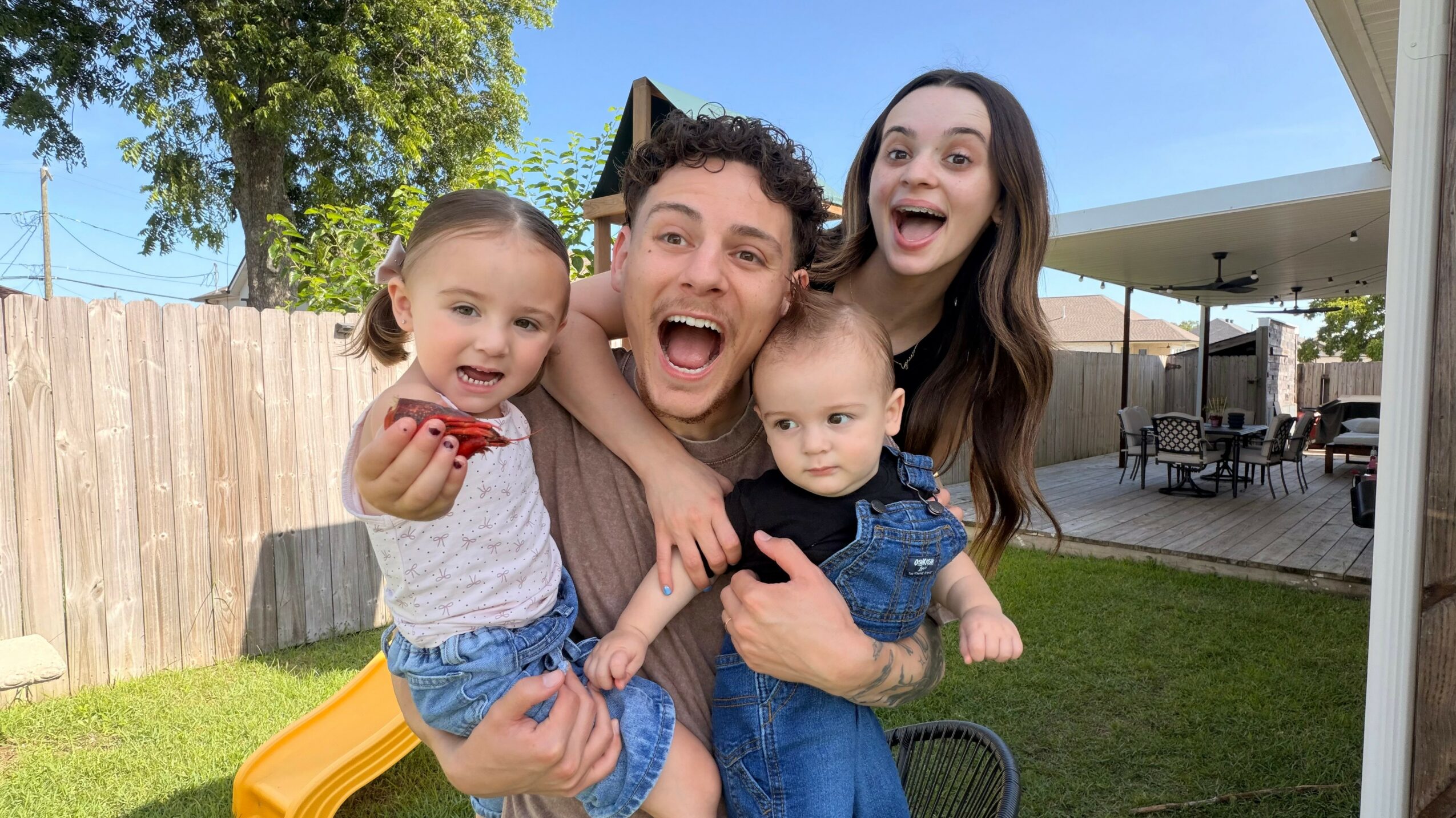 Family of four smiling outdoors in a backyard, with a wooden fence and patio in the background.