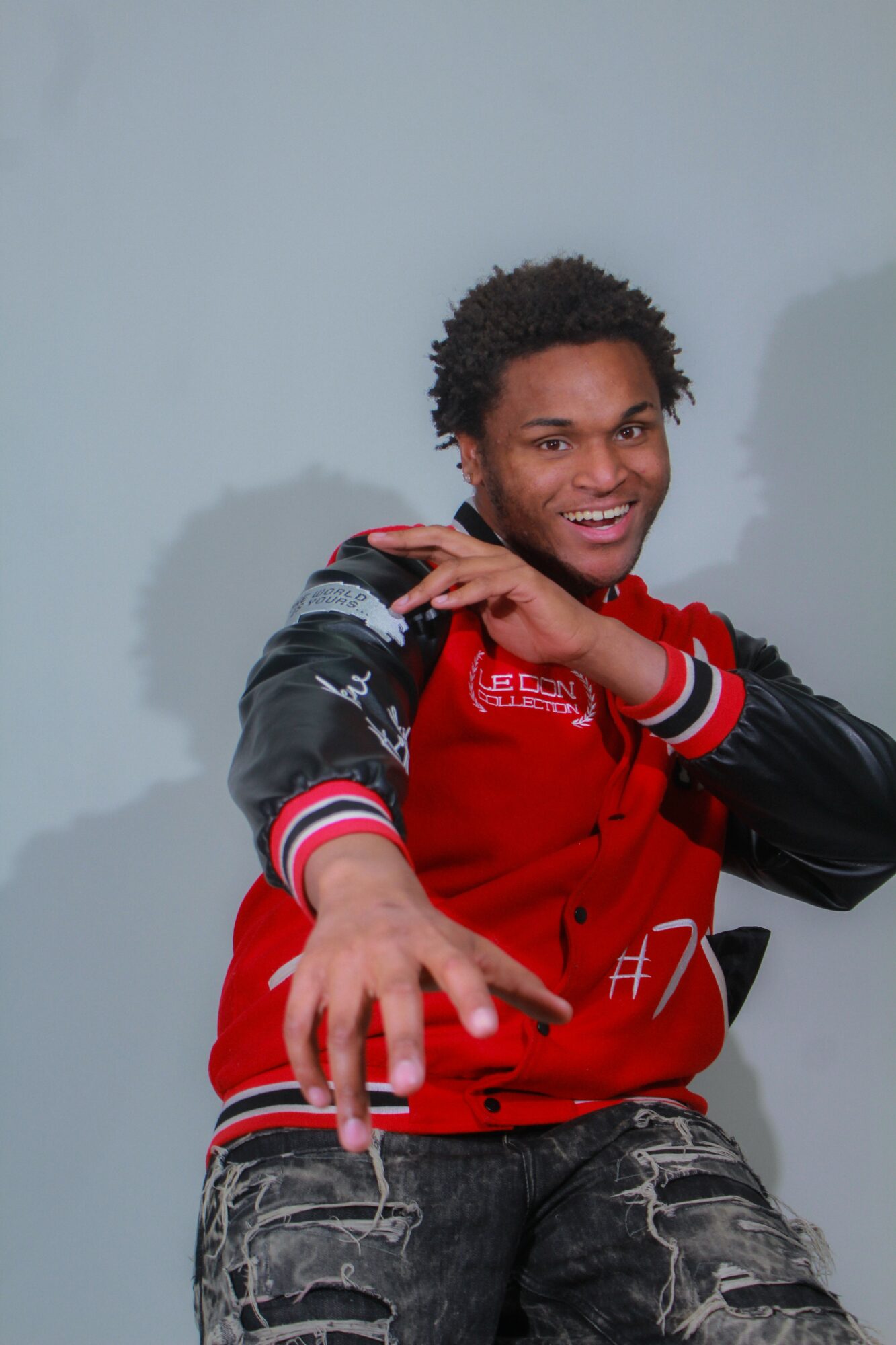 Young man with curly hair smiling, wearing a red and black jacket, reaching forward with his hand, against a plain background.