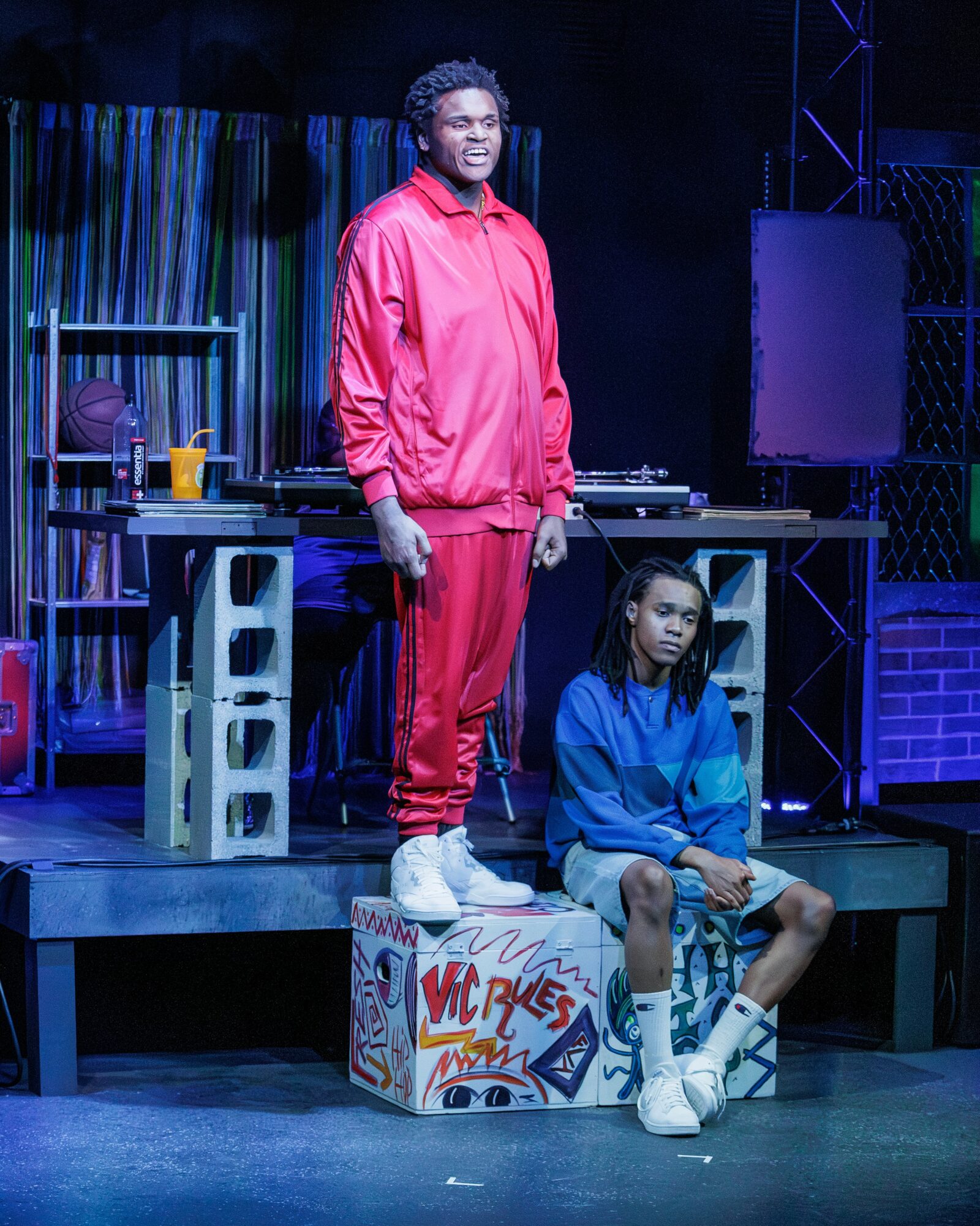 Two people on stage, one standing in a red tracksuit, the other sitting in a blue outfit, with stage props and equipment.