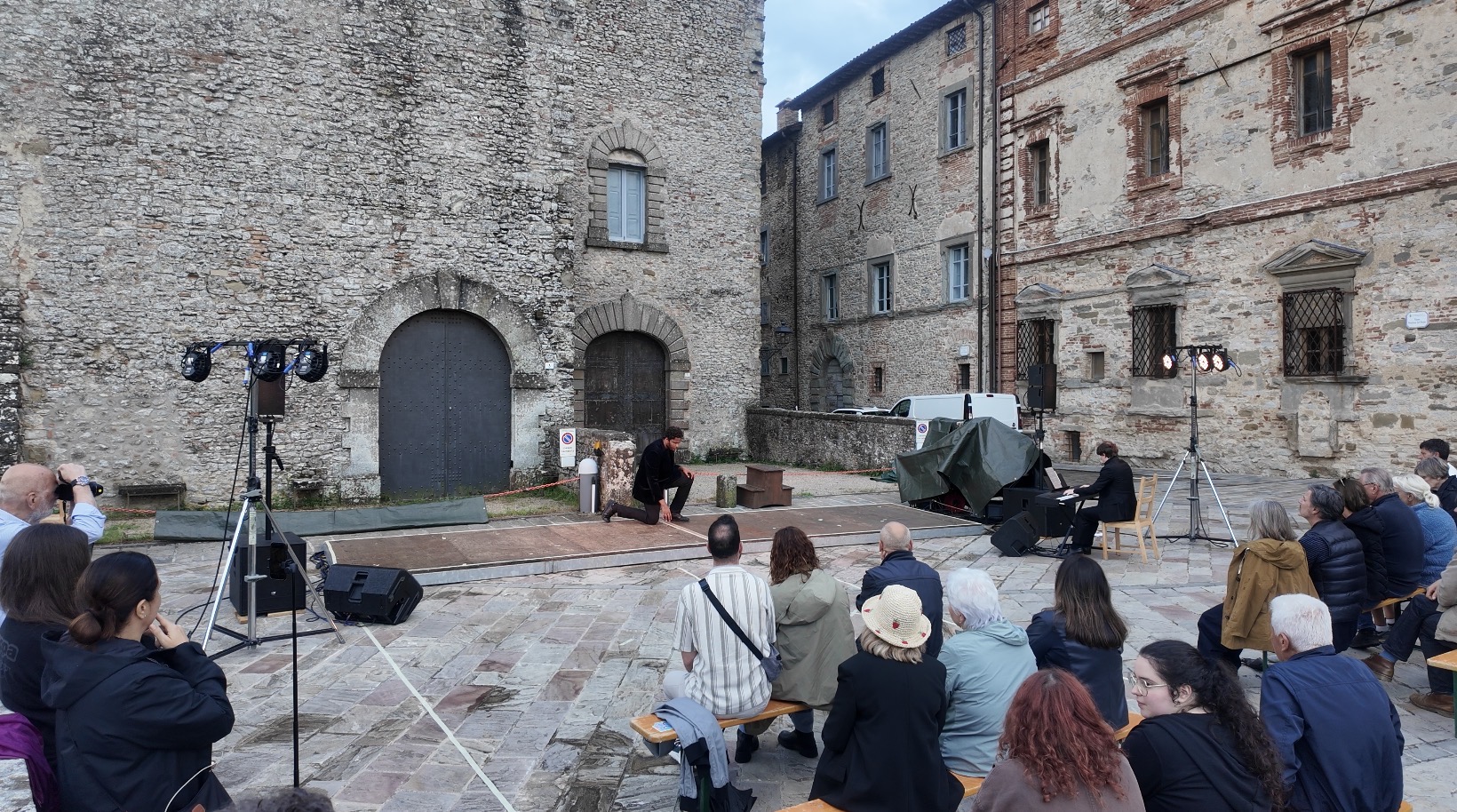 People seated outdoors in front of historic stone buildings, some with military vehicles nearby, during daytime.