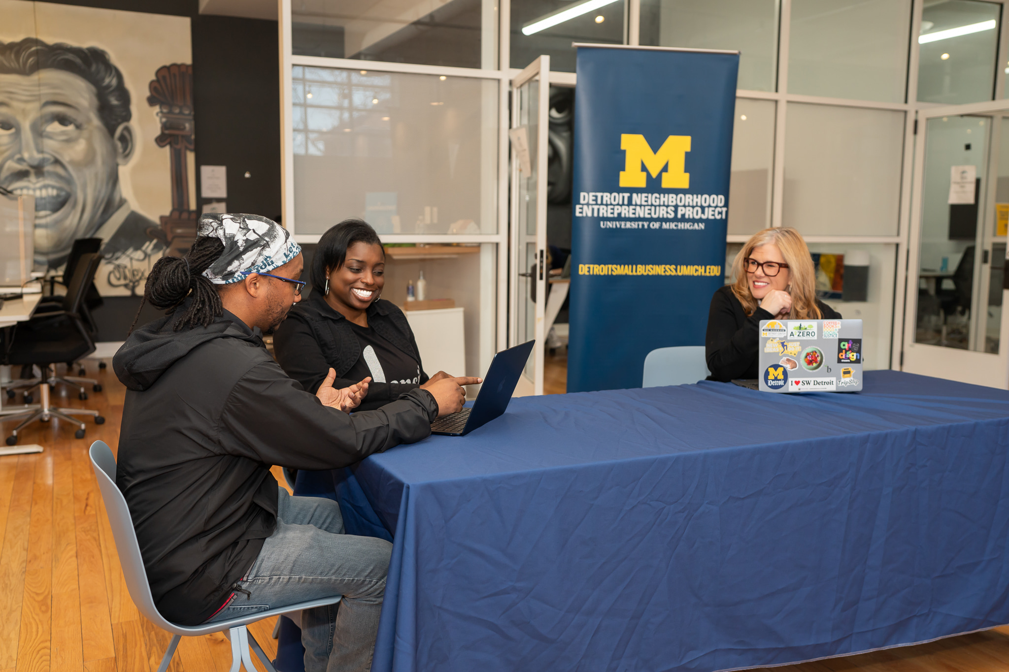 Three people sitting at a table with a University of Michigan banner behind them, one person using a laptop, two smiling.