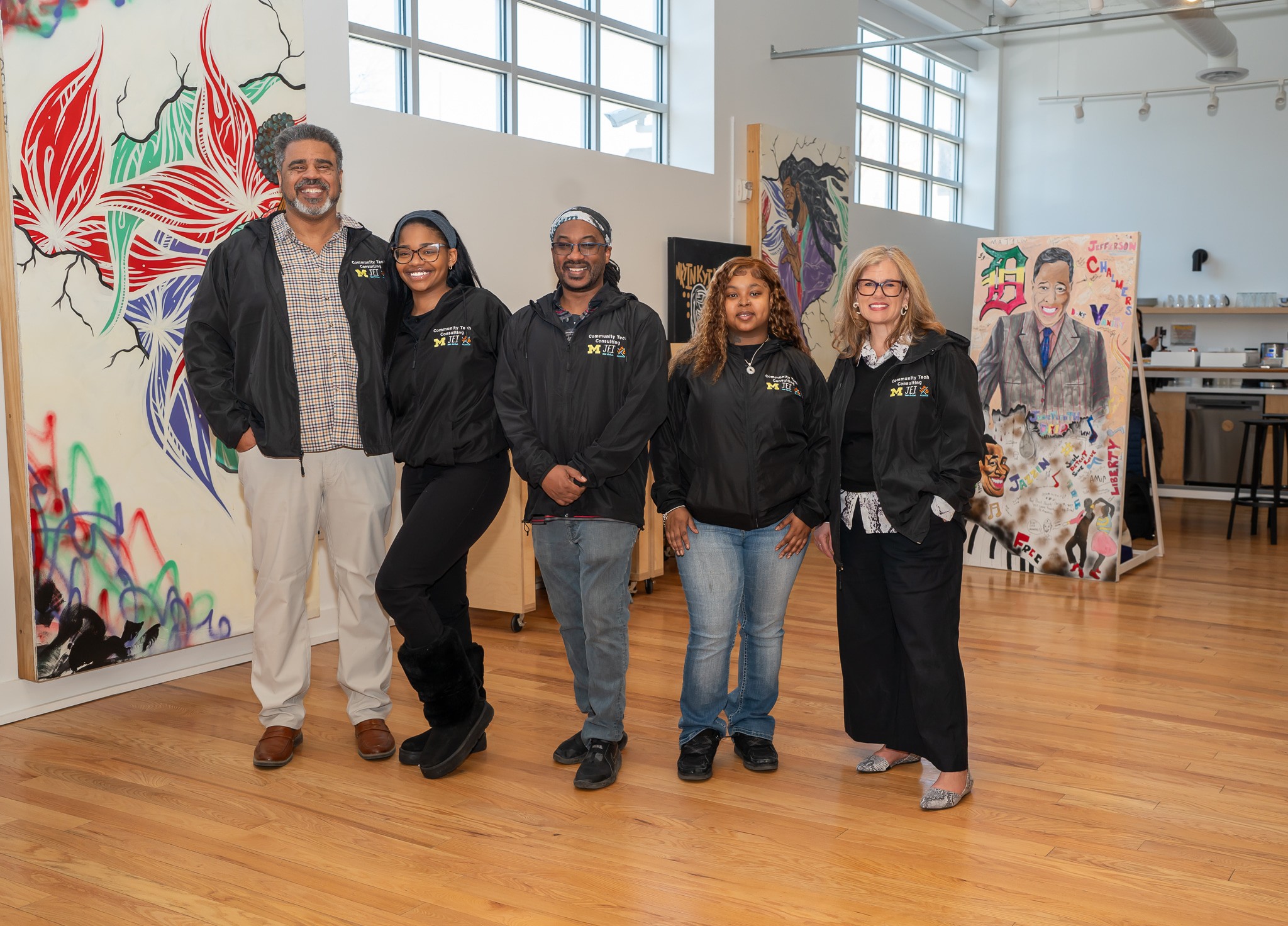 Group of five people standing in an art studio with colorful paintings and large windows, smiling at the camera.