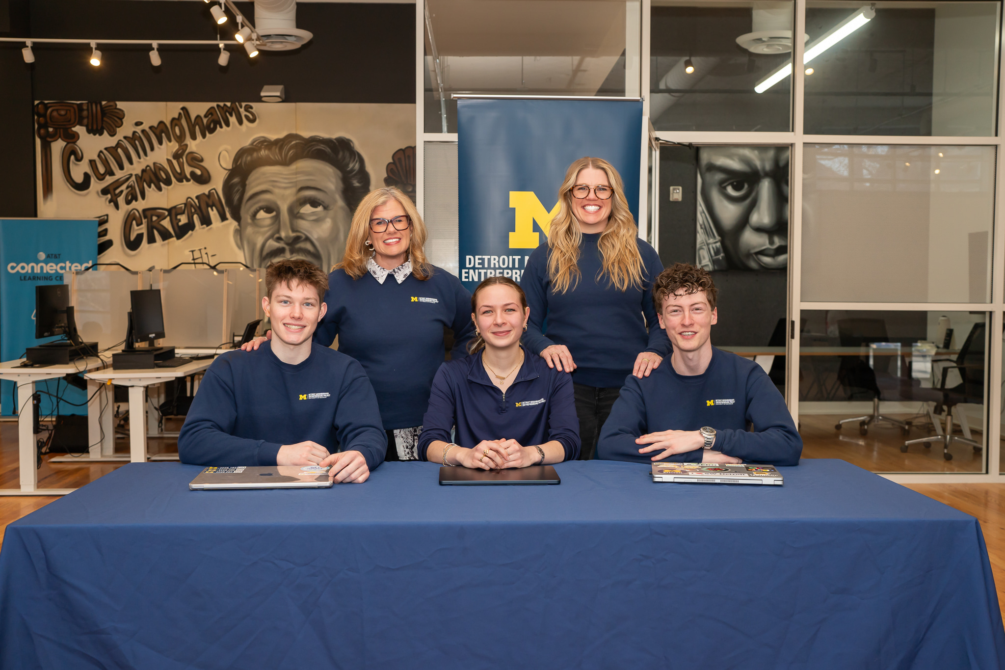 Five people in navy uniforms sit and stand behind a table with a blue tablecloth, in a modern room with windows and posters.