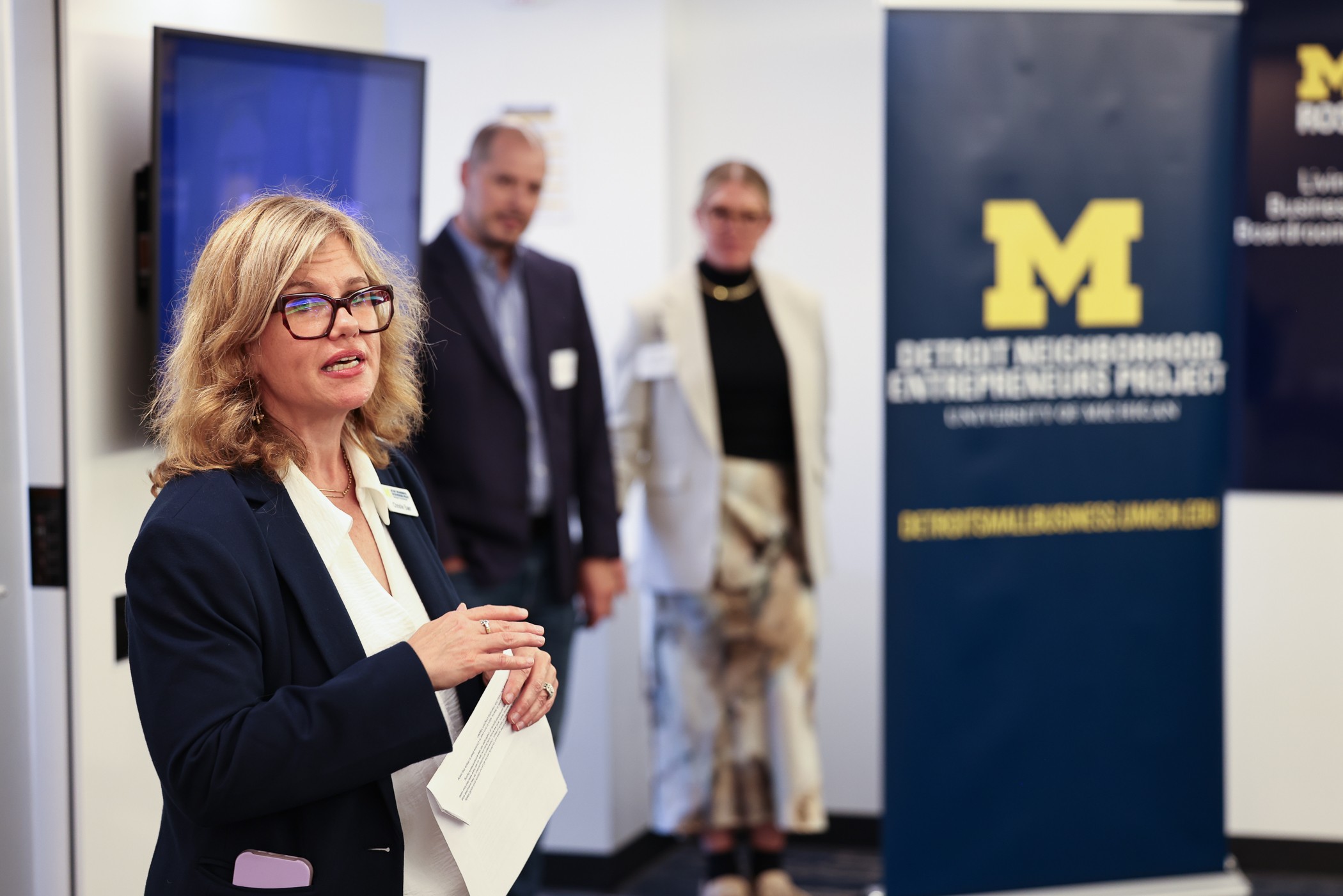 Woman with glasses speaking at an event, with three people and a University of Michigan banner in background.