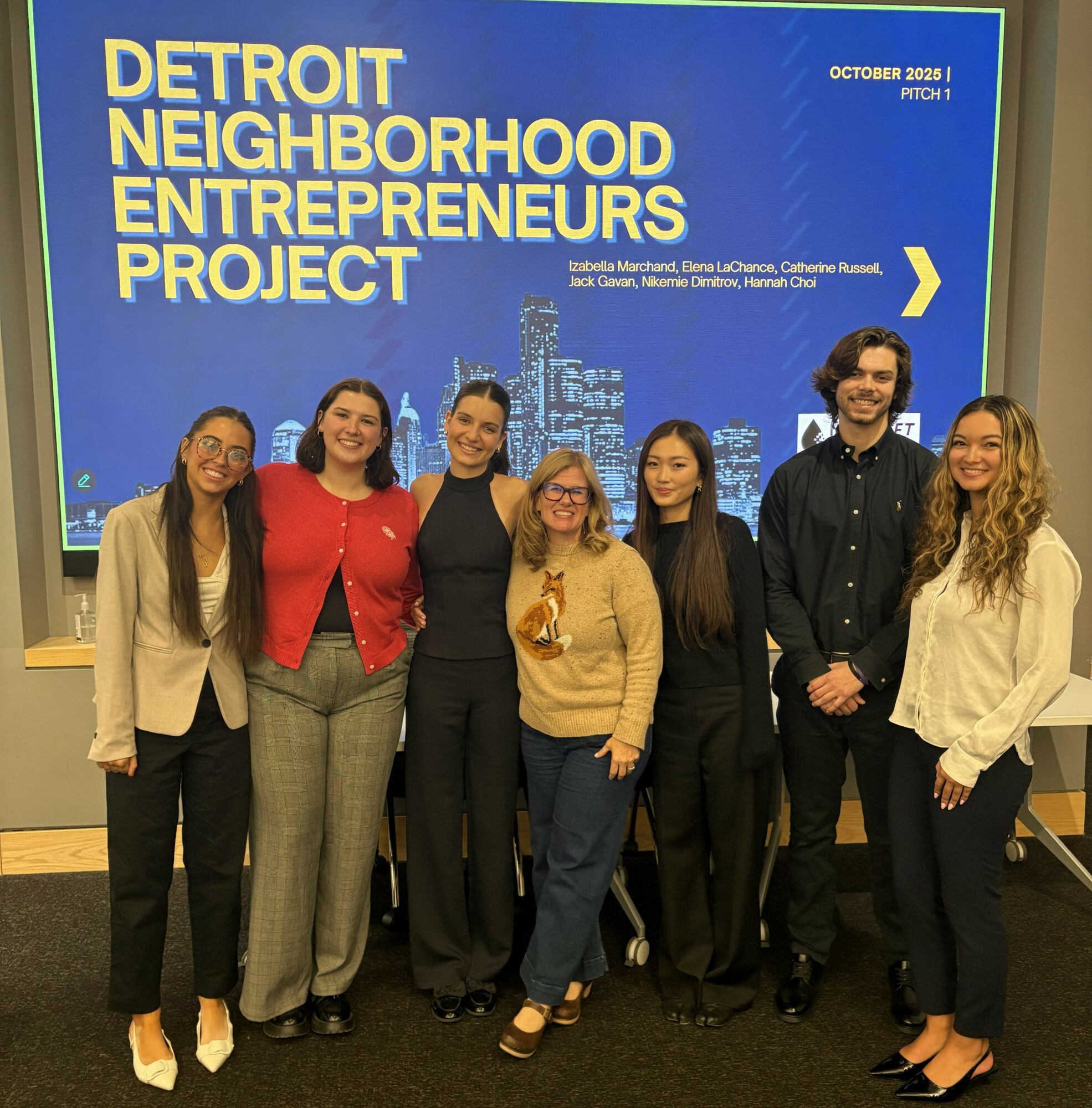 Group of seven people standing in front of a blue presentation screen with text about Detroit Neighborhood Entrepreneurs Project, October 2023.