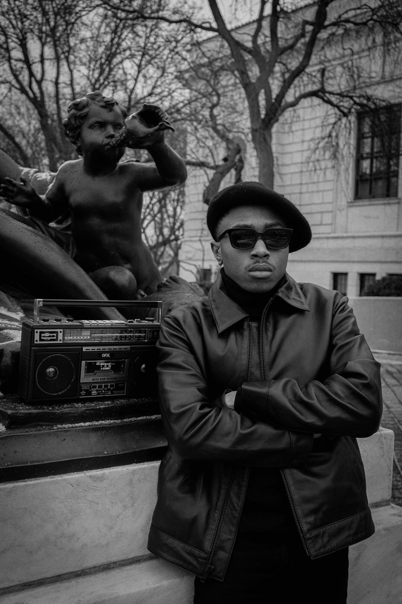 A man wearing sunglasses and a hat stands with arms crossed in front of a building, with a child sitting on a car behind him, holding a bottle.
