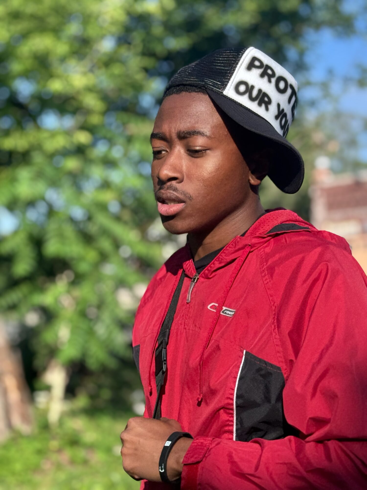 Young man wearing a red jacket and black cap outdoors, looking to the side with trees in background.