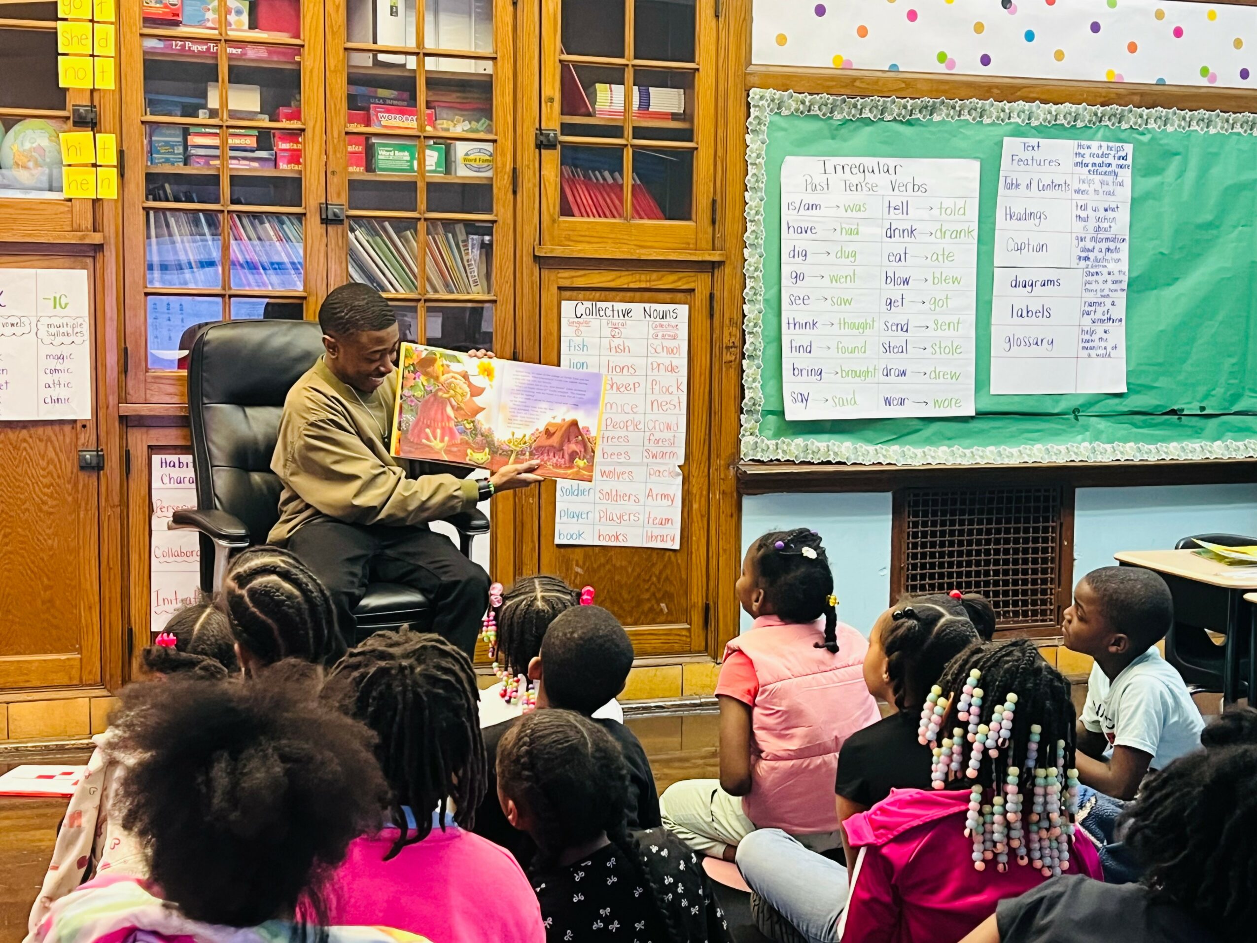 Adult reading a colorful book to children seated on the floor in a classroom with bookshelves and a green bulletin board.