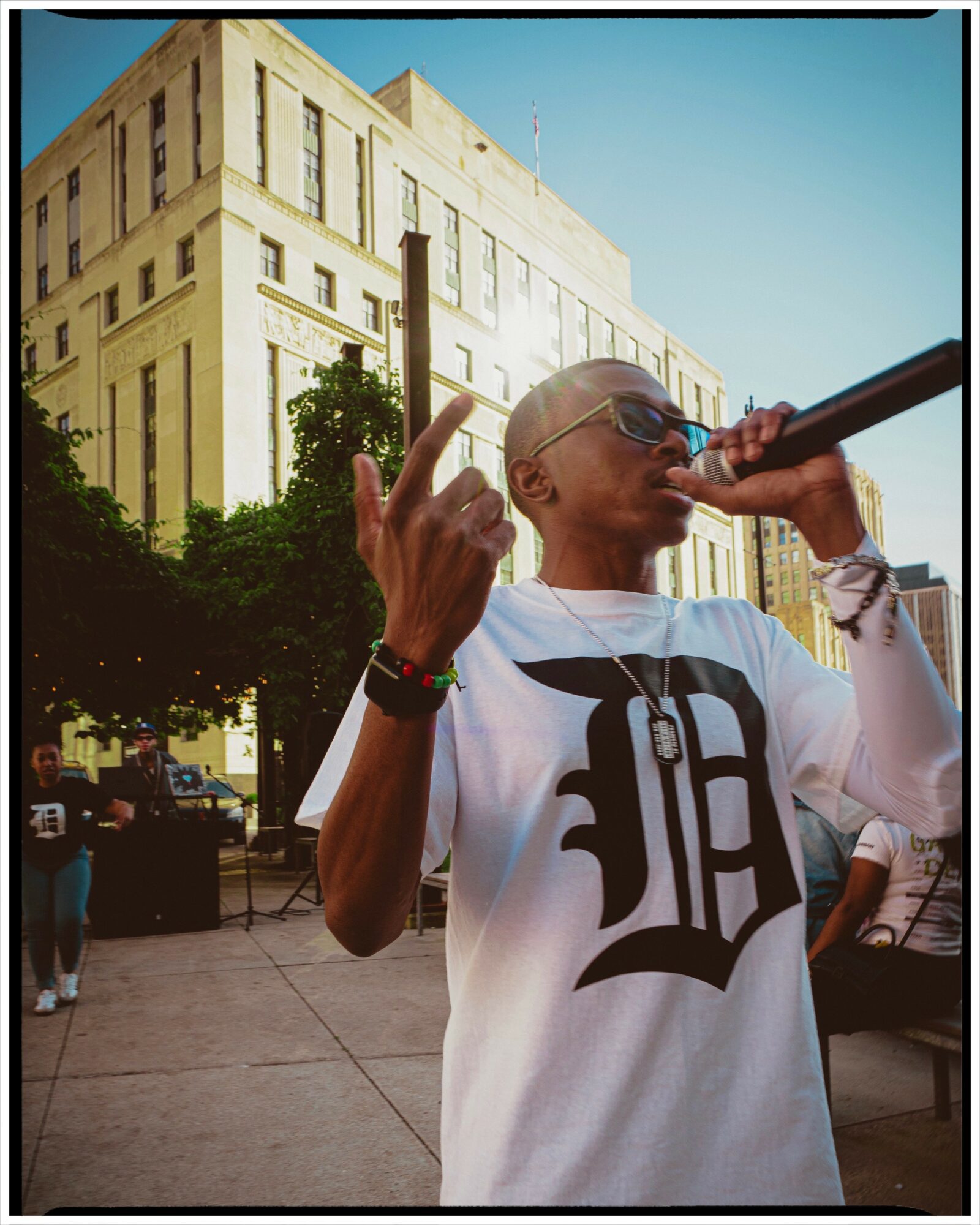 Person with sunglasses and a white shirt holding a microphone outdoors, with a large building and trees in the background.