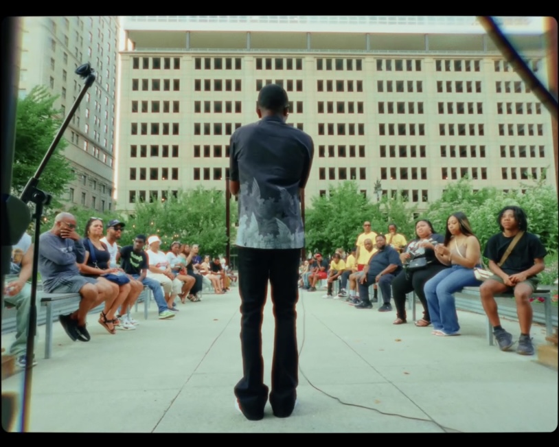 Person standing with back facing camera, audience seated in a semi-circle outdoors, large building in background.