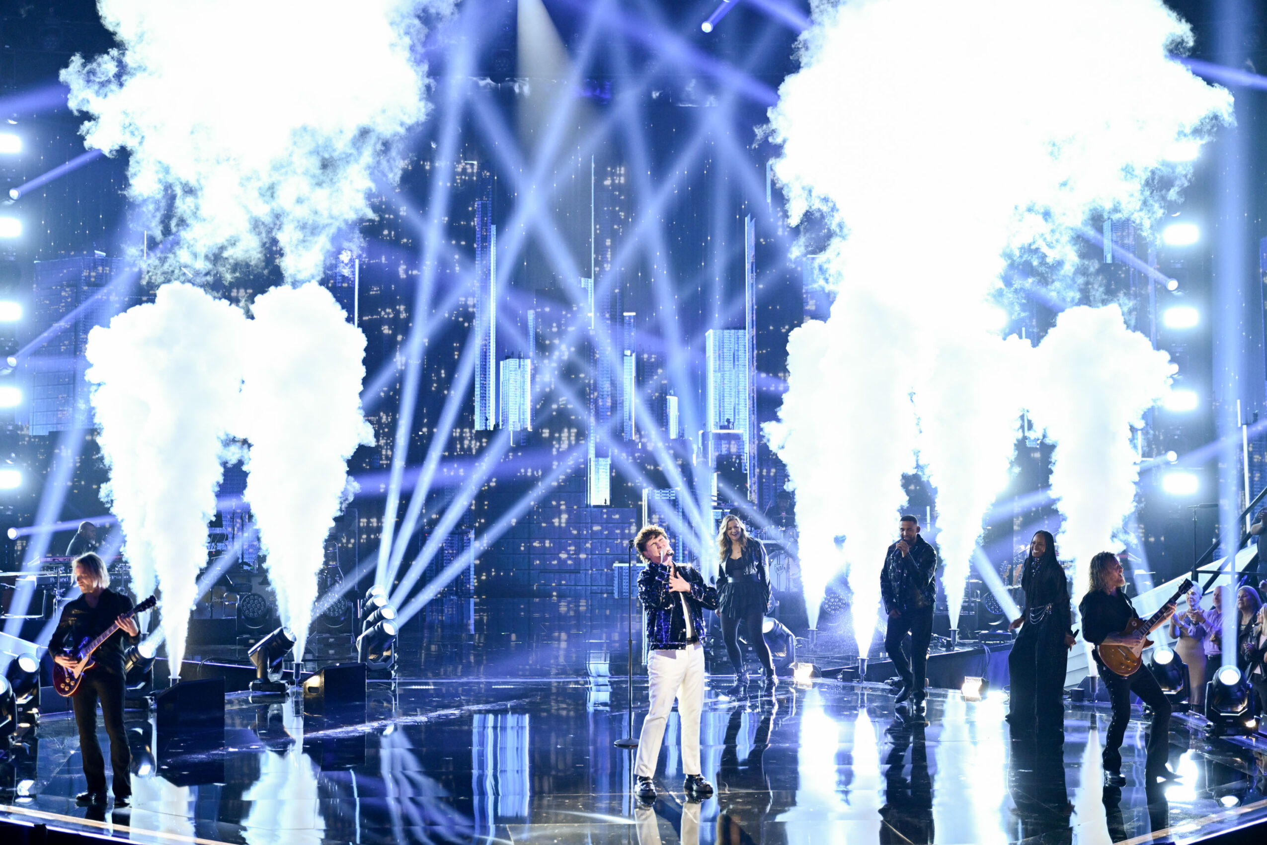 Musicians perform on stage with bright lights, smoke effects, and laser beams, creating a lively concert atmosphere.