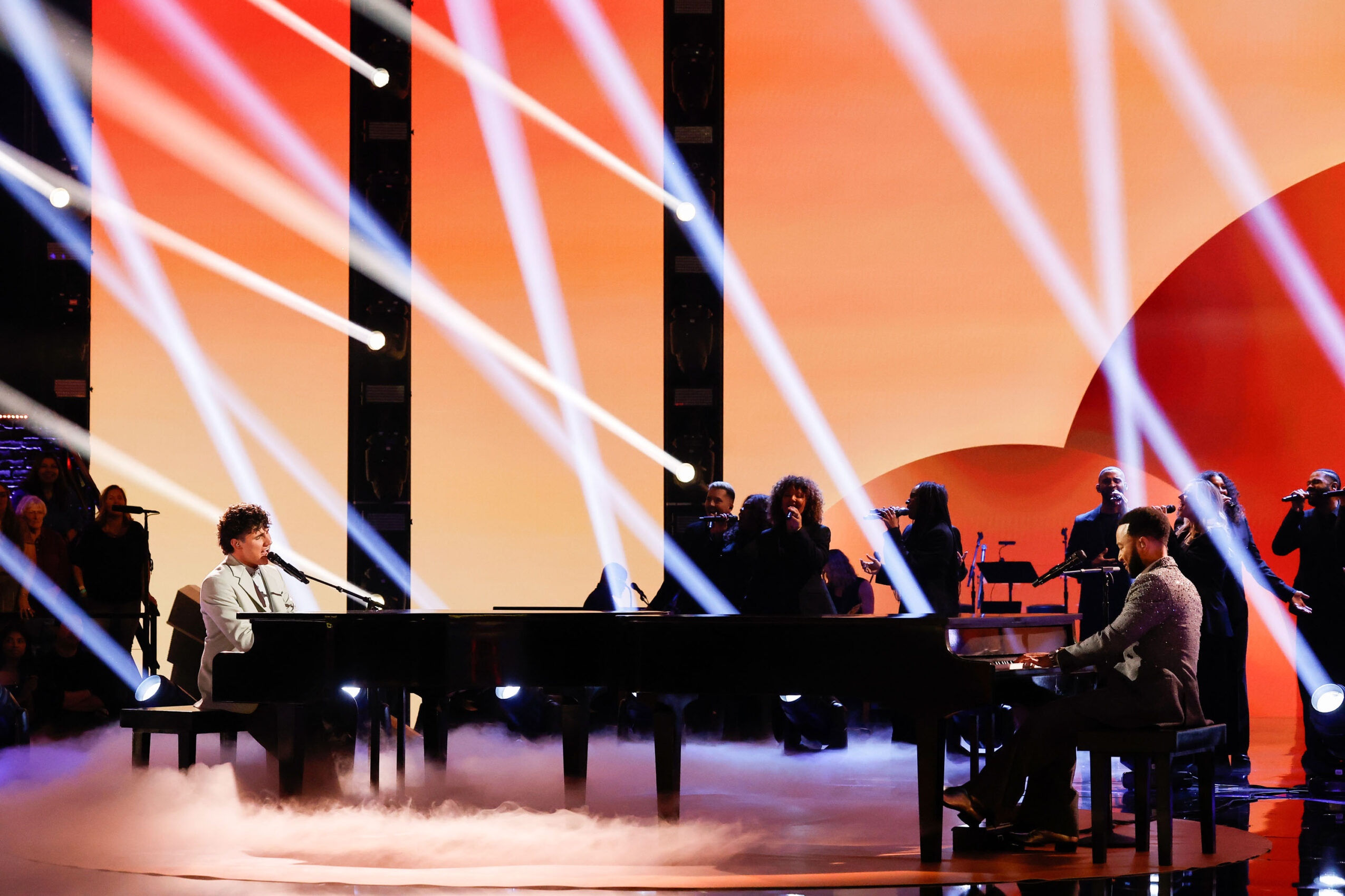Musicians perform on stage with bright lights and a sunset backdrop, playing pianos and other instruments.