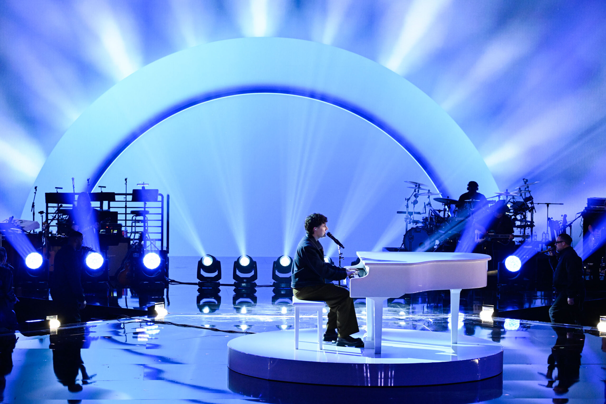 Person playing white piano on stage with blue lighting and circular backdrop, surrounded by stage equipment.