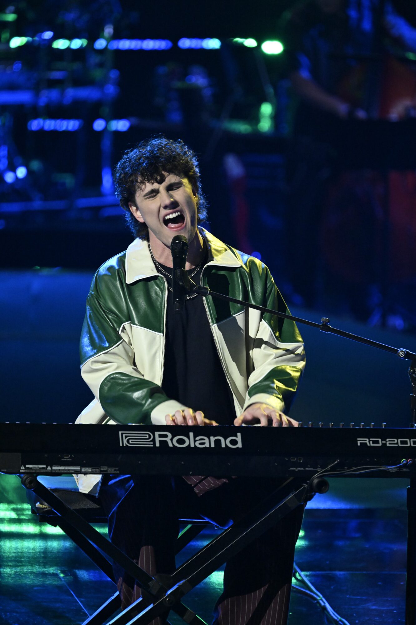 Person singing passionately while playing a keyboard on stage with blue and green lighting