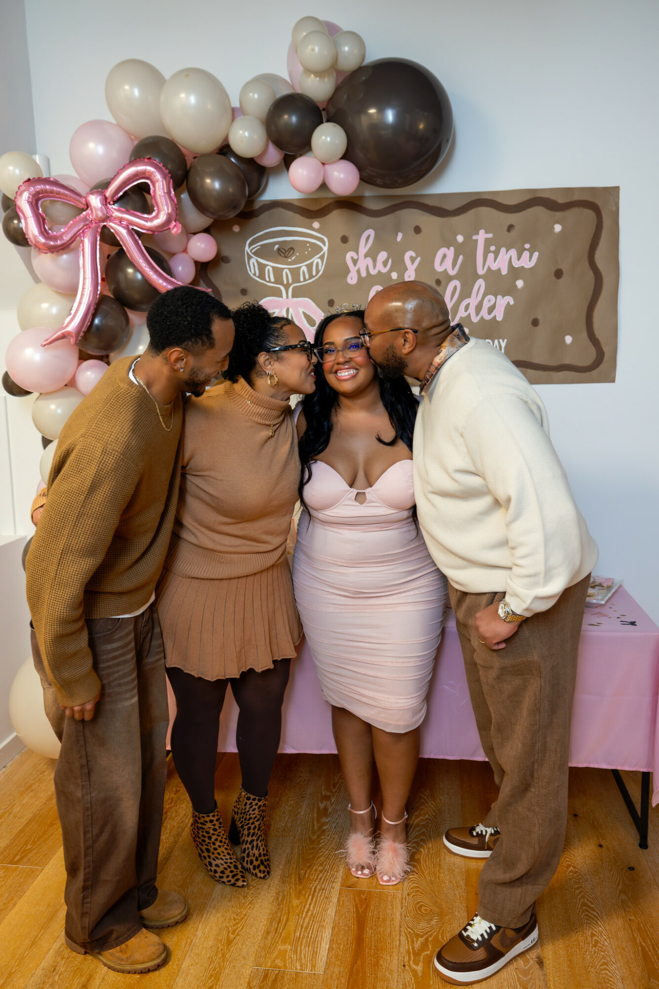 Four people smiling and posing together in front of balloon decorations and a sign, indoors.