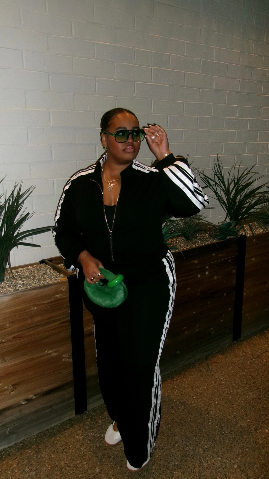 Person in black tracksuit with white stripes, sunglasses, holding a green purse, standing outdoors near plants and a white brick wall.