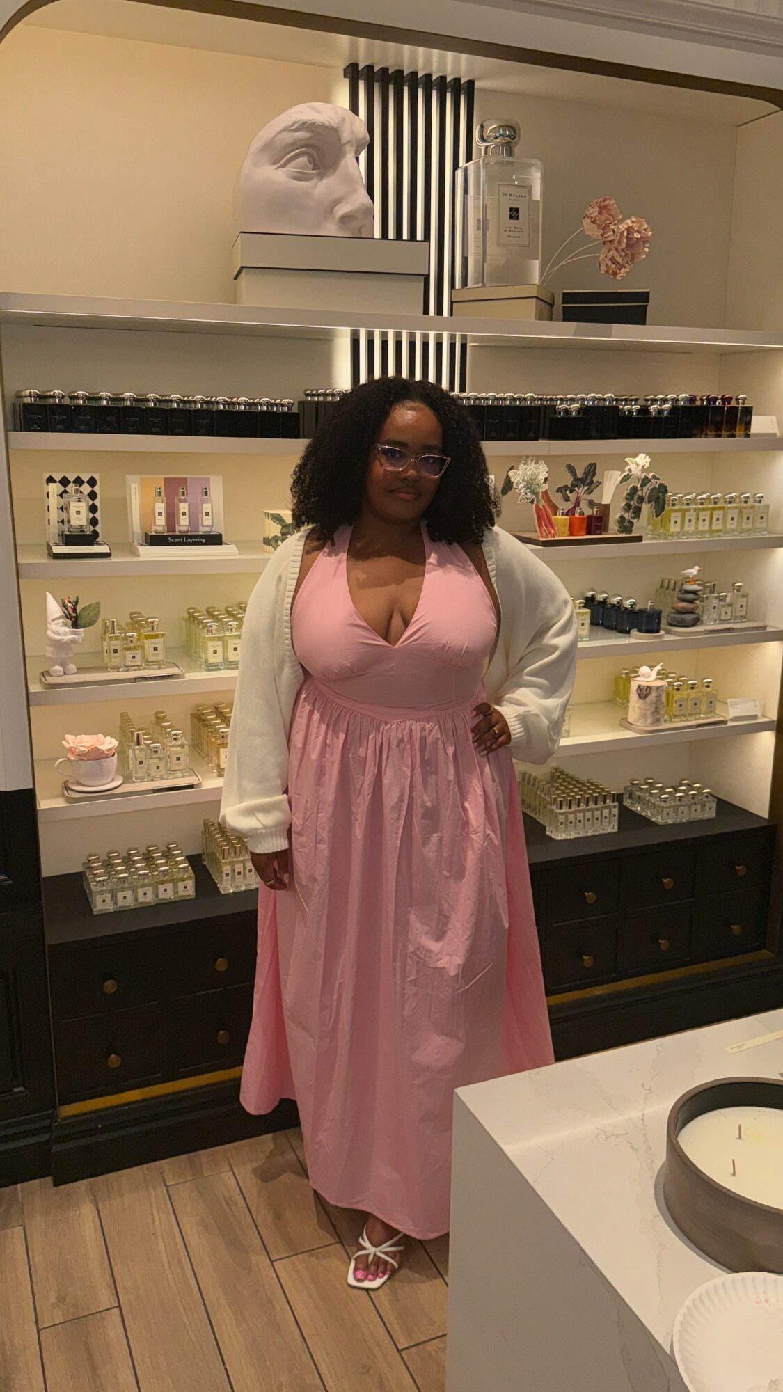 Woman in pink dress and white cardigan standing in front of shelves with decorative items, smiling, in a store or showroom.