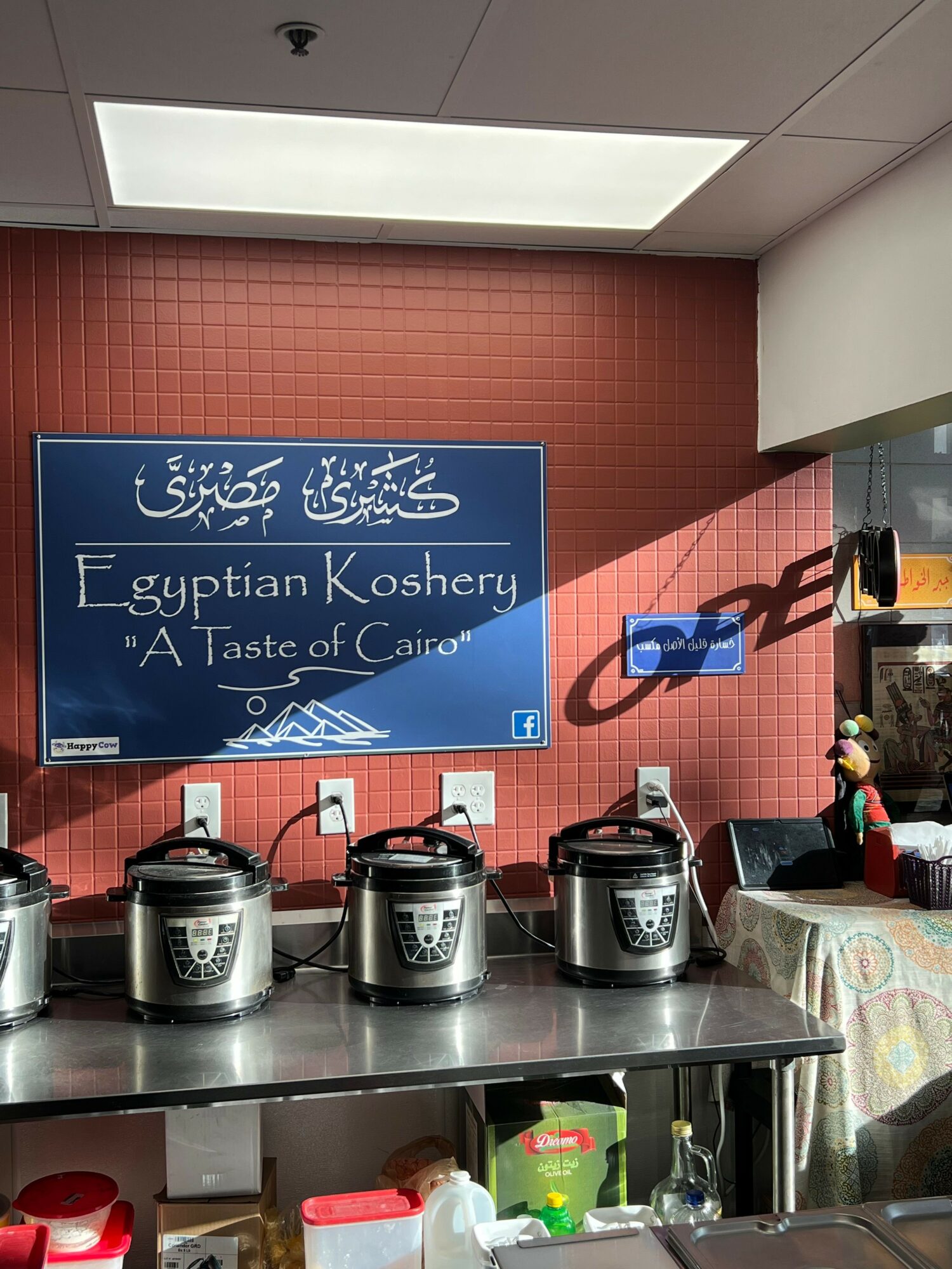 Coffee shop interior with a blue sign displaying Arabic and English text, and coffee machines on a counter.