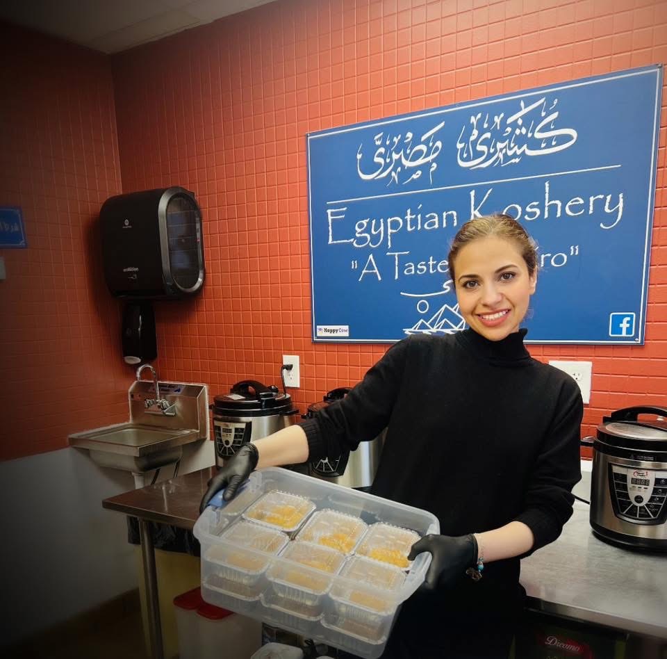 Woman in black sweater holding a container of food in a kitchen with a blue sign and kitchen appliances.