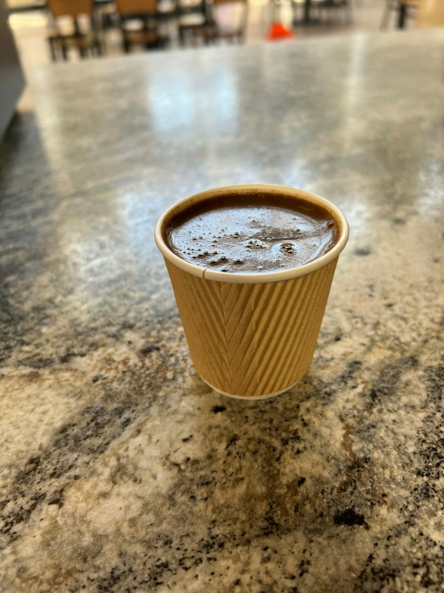 A disposable coffee cup filled with a hot beverage on a textured surface, with foam on top.